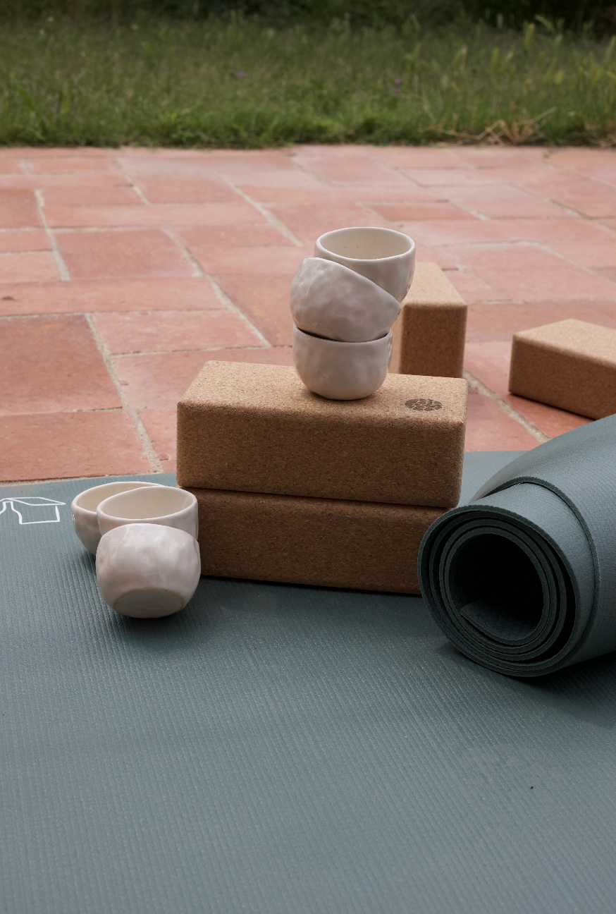 Tapis de yoga, blocs en liège, tasses en céramique, et rouleau de yoga sur une surface extérieure en briques, avec de l'herbe en arrière-plan.