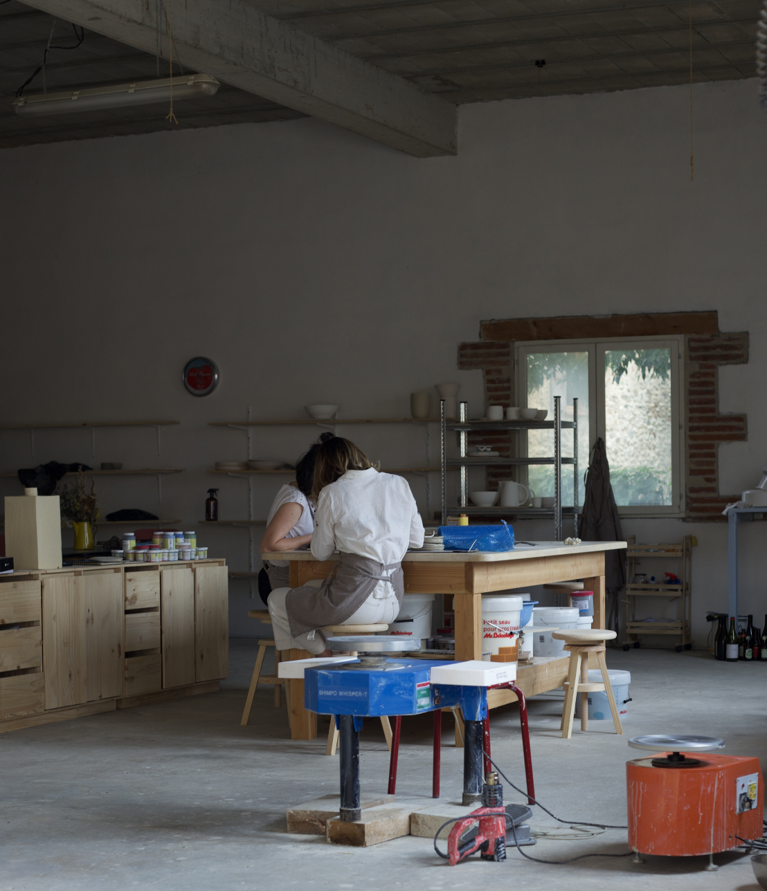 Deux personnes travaillent ensemble à une table dans un atelier avec des étagères et des ustensiles, l'une d'elles porte un tablier