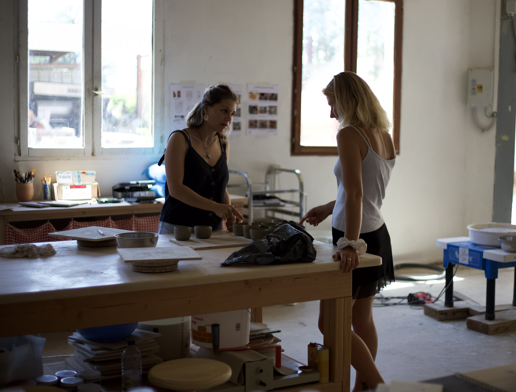 Deux femmes discutent dans un atelier ou une pièce de travail, entourées de poteries et d'outils.
