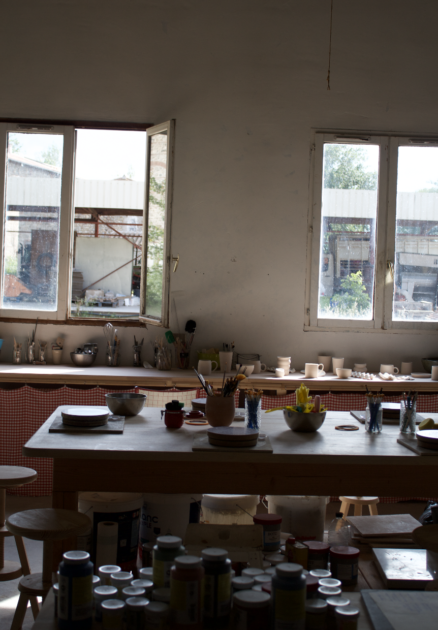 Intérieur d'un atelier avec une grande table en bois, de nombreux bacs, pinceaux, tasses, et pots de peinture, lumières naturelles entrant par deux fenêtres, vue extérieure d'une zone de travaux ou stockage