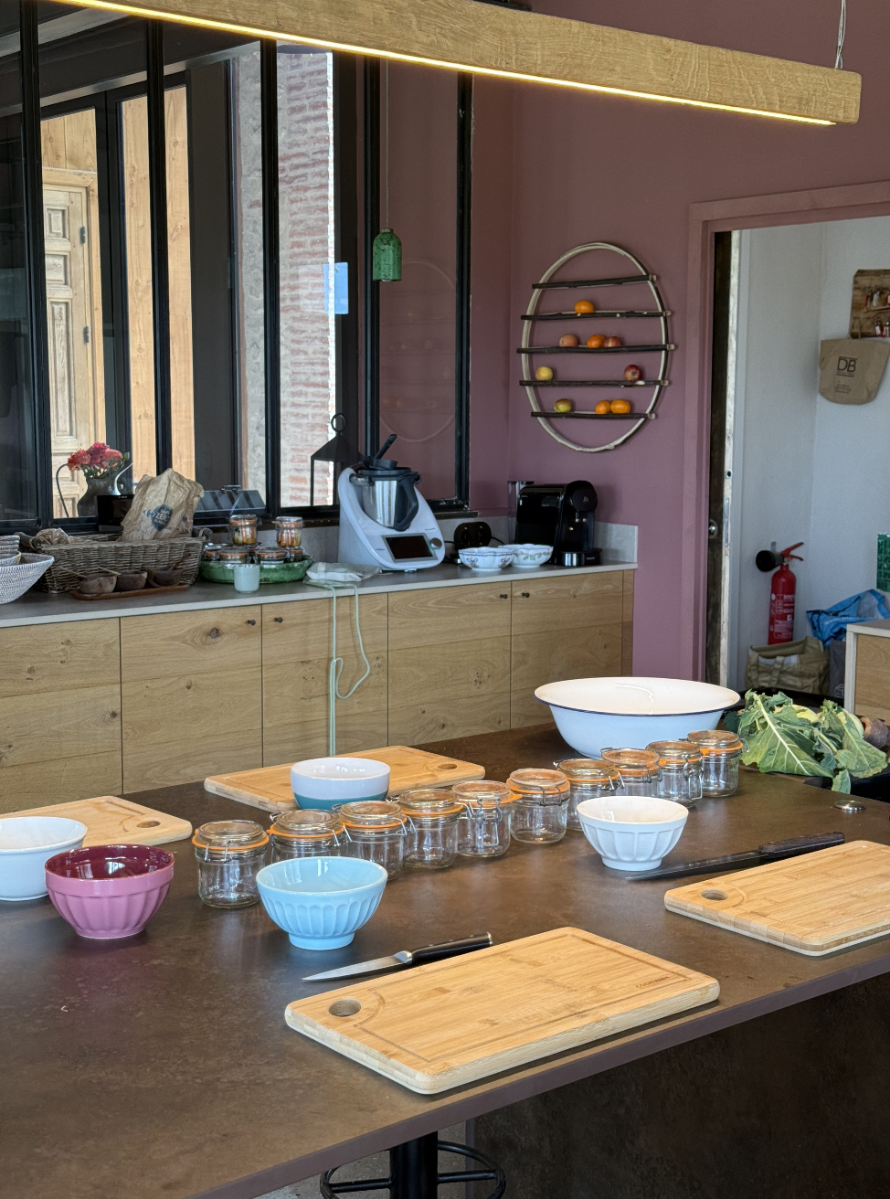 Cuisine avec une table en bois, plusieurs bocaux en verre, bols en céramique colorés, planches à découper, et une partie de la cuisine avec des appareils électroménagers, une étagère à fruits murale, et une fenêtre.