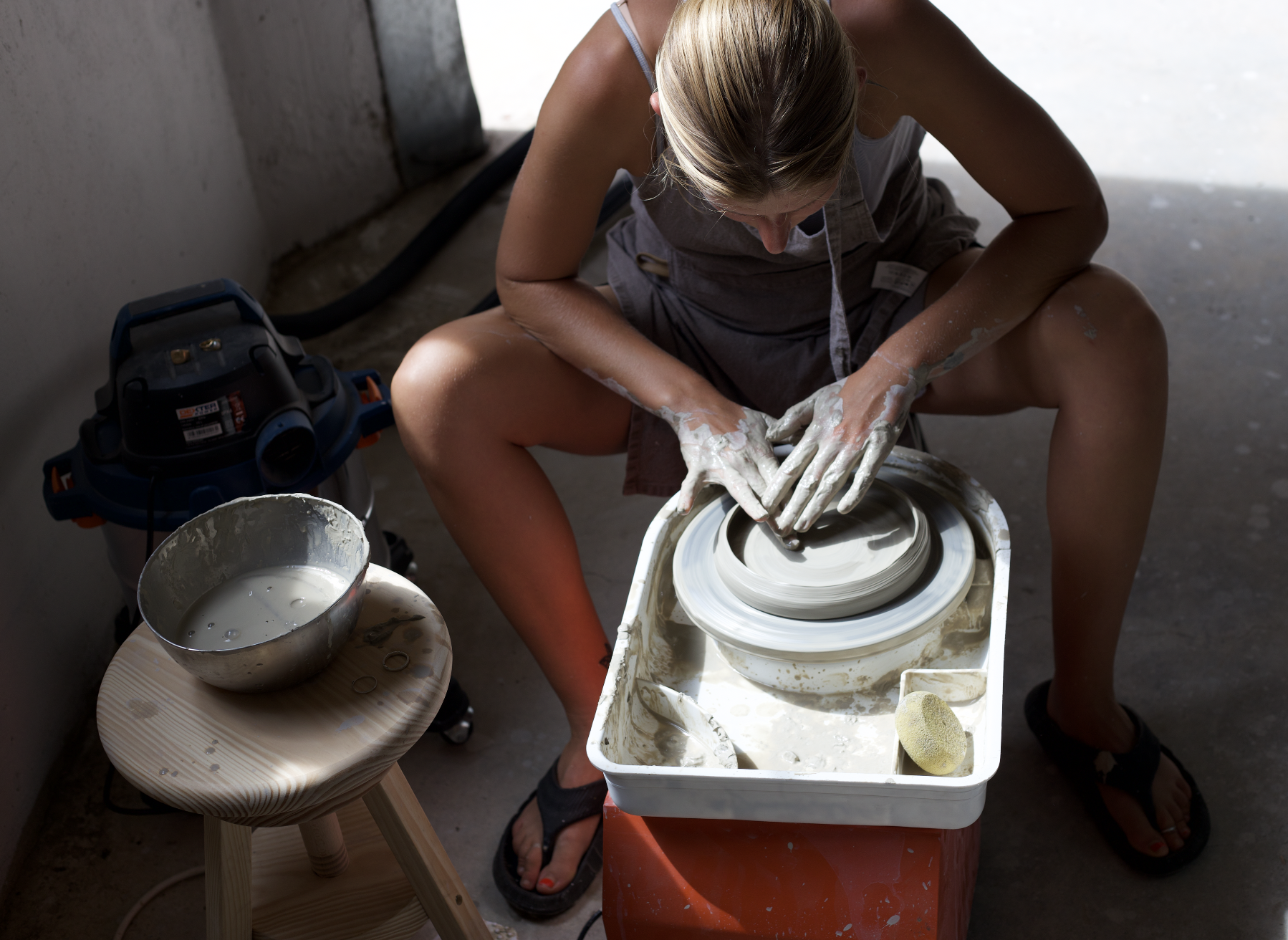 Femme moulant de la poterie sur un tour de poterie, utilisant ses mains pour façonner l'argile, avec un seau d'eau et un tampon à éponges à proximité.