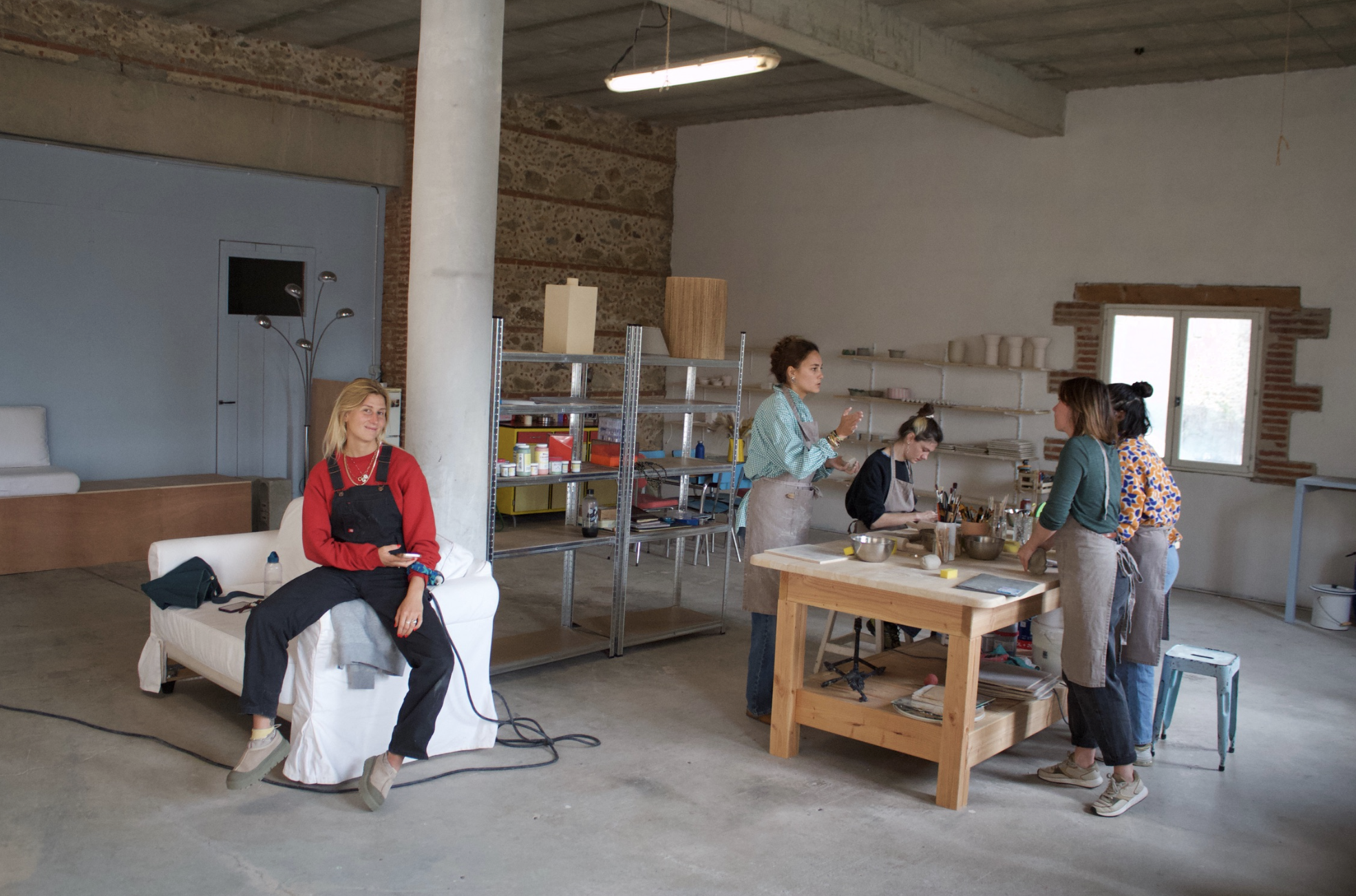 Une femme assise sur un canapé blanche, souriante, dans un studio de cuisine, avec un groupe de quatre femmes en train de cuisiner à une table en bois.