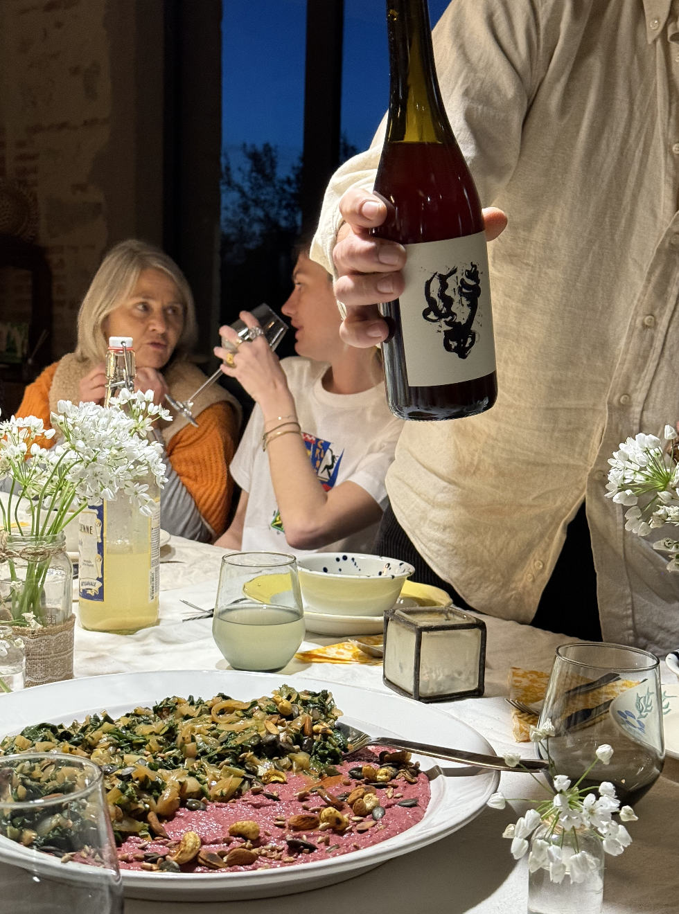 Une table dressée pour un dîner avec un plat de viande rosée garnie de légumes et d'oléagineux, des verres de boissons, des fleurs en décoration, et des personnes discutant dans une ambiance chaleureuse.