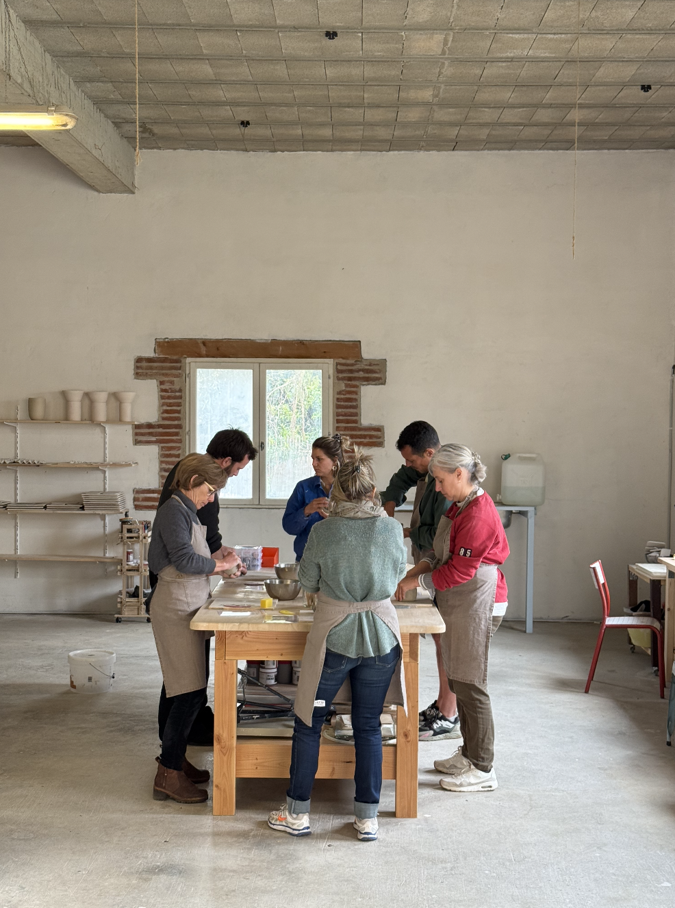 Groupe de personnes en train de cuisiner ou de préparer quelque chose sur une grande table en bois dans un atelier ou une cuisine, dans une salle avec un mur blanc, une fenêtre, et une étagère avec des pots et des assiettes.