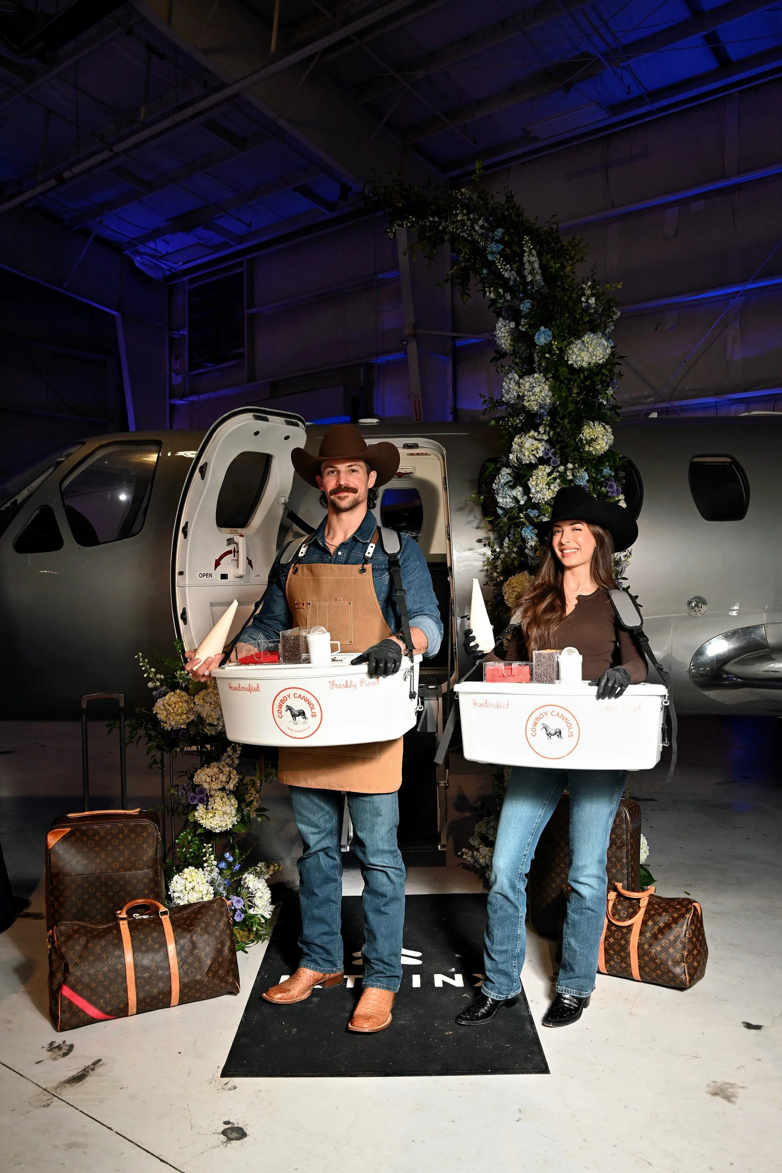Two people dressed as cowboys standing in front of a private jet, holding boxes of charcuterie. They are smiling and wearing cowboy hats and gloves. Flowers and luggage are around them, with a large floral arrangement on the jet.