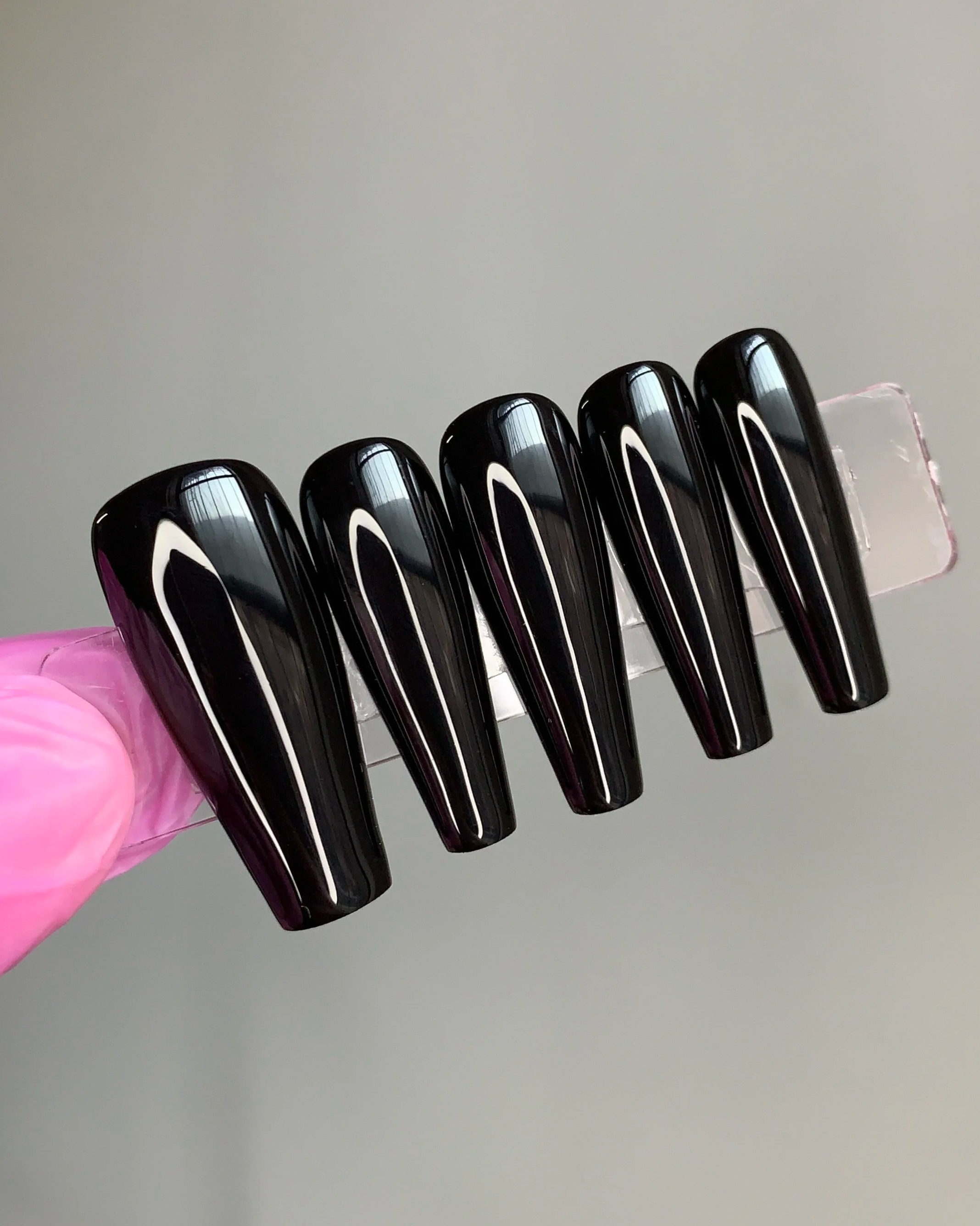 Long coffin press-on nails on a clear strip held in front of a grey wall.