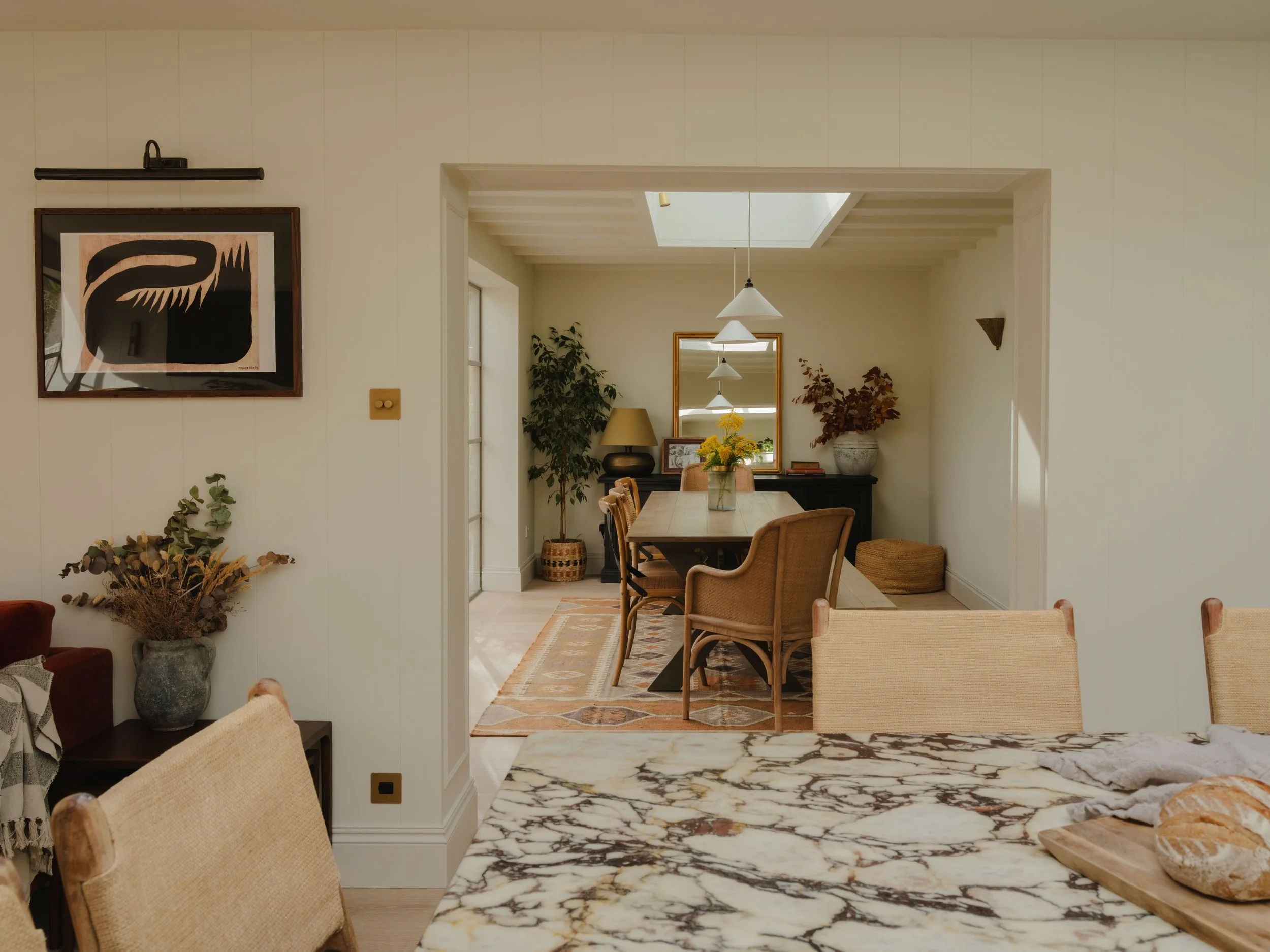 A view from a kitchen looking into a dining area with a wooden table, chairs, a mirror, plants, and hanging pendant lights.