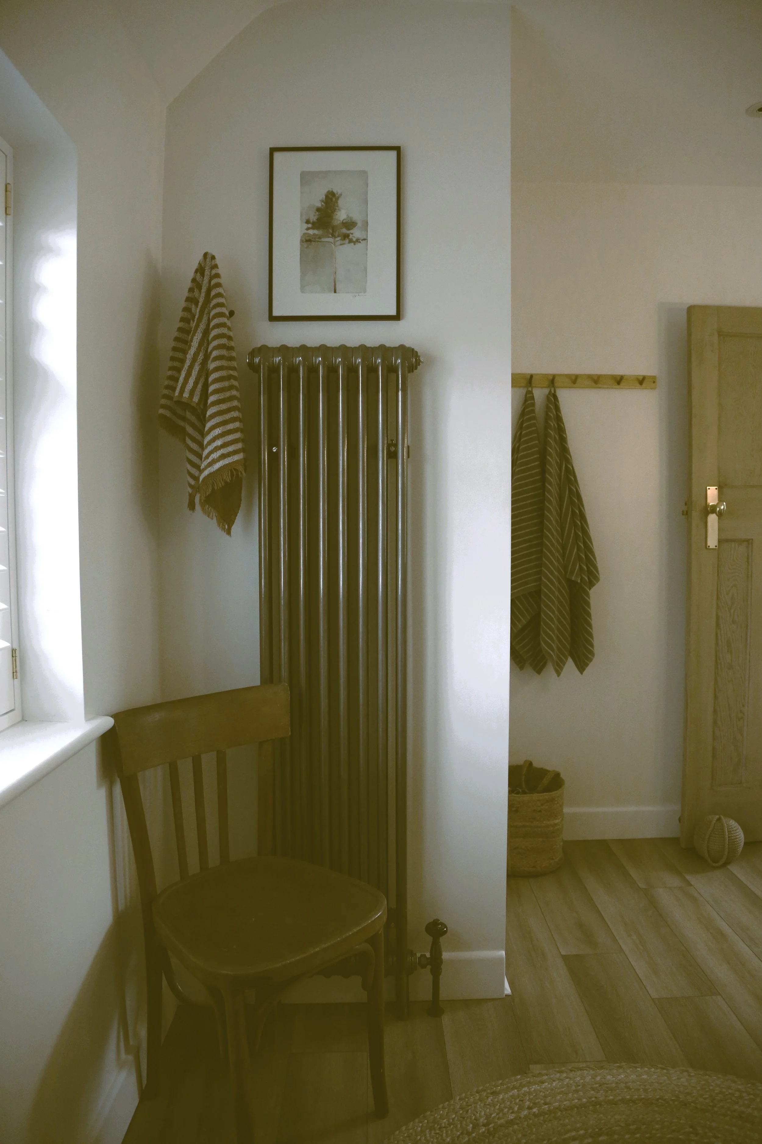 A cozy corner of a room with a wooden chair, a window with shutters, striped towels hanging on a wall, a tall vintage radiator, framed art on the wall, and a doorway with a basket and a crochet ball nearby.
