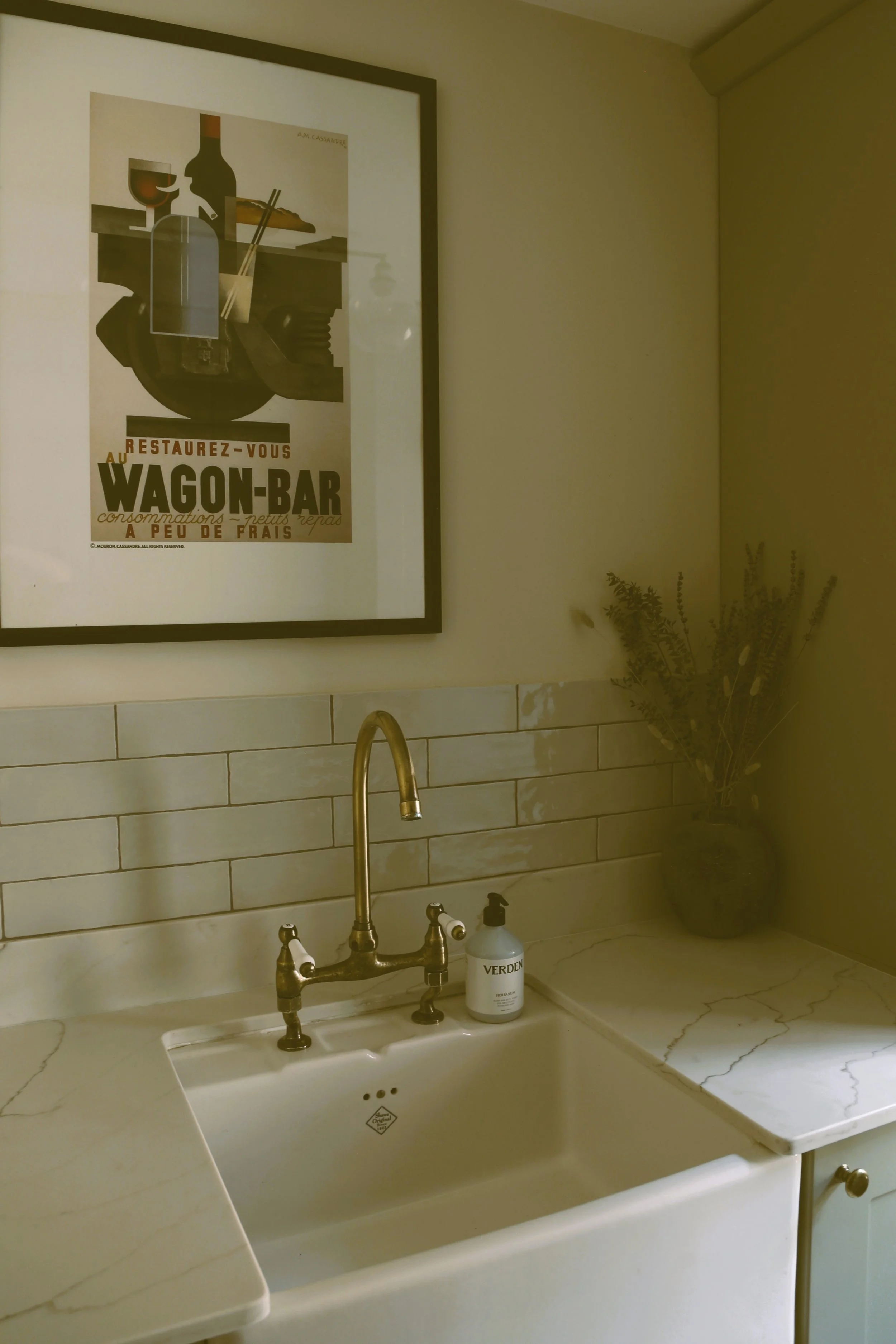A kitchen sink with brass faucet, soap dispenser labeled 'VERDEEN,' a dark vase with dried plants, a framed vintage-style poster, marble countertop, and cream-colored tiled backsplash.