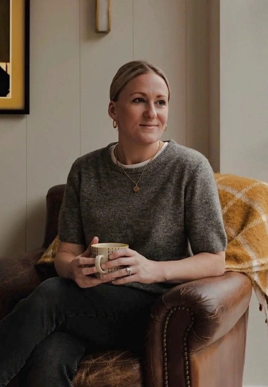 A woman sitting on a leather couch, holding a mug, with a yellow and brown blanket draped over the armrest, in a cozy room with light-colored walls and a framed picture on the wall.