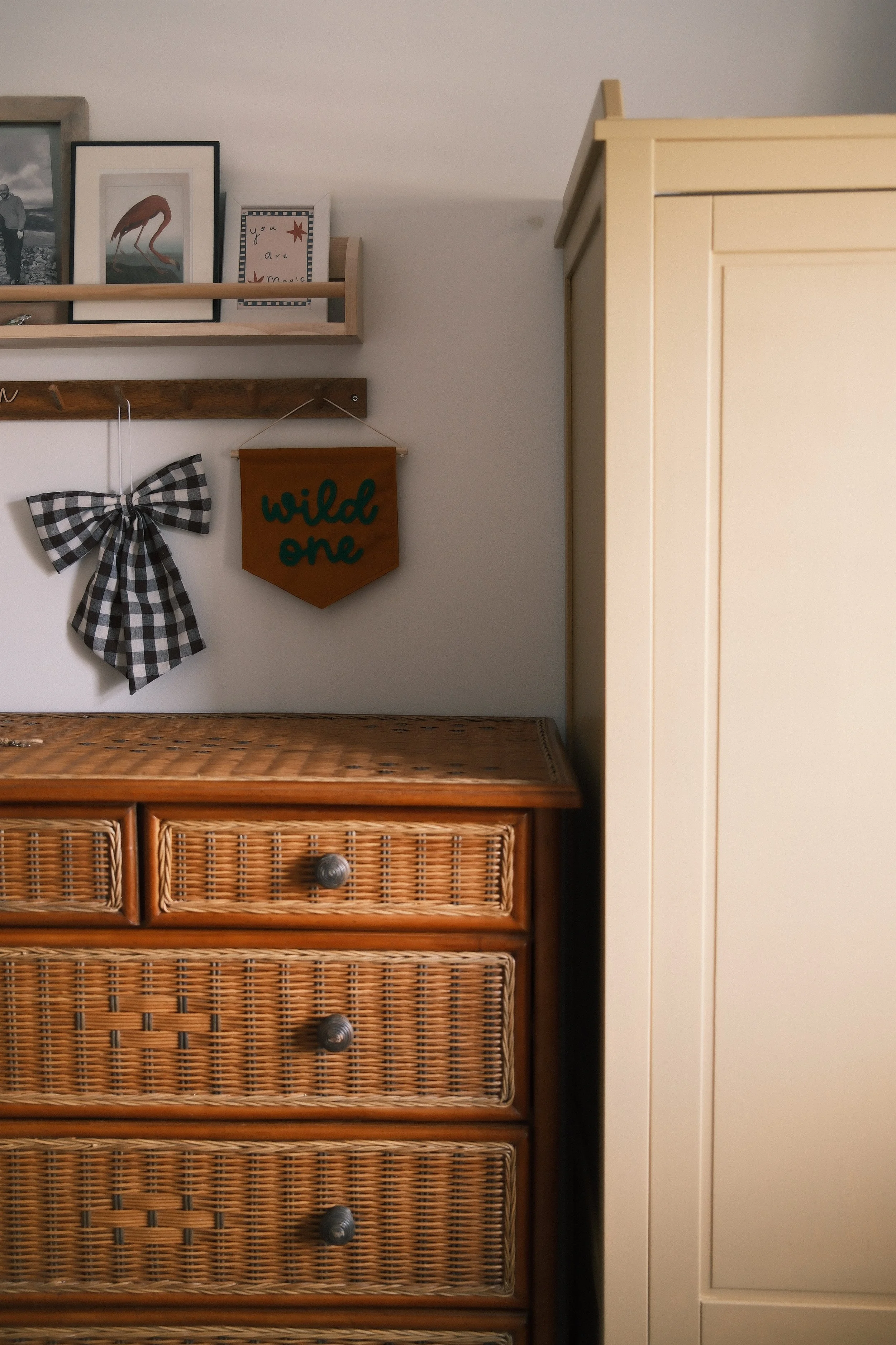 Wooden dresser with woven rattan drawers and a dark wood sign that says "wild one" hanging on the wall next to it. Above, a wooden shelf with framed pictures and a small decorative sign that says "you are magic." A black and white checkered bow decor