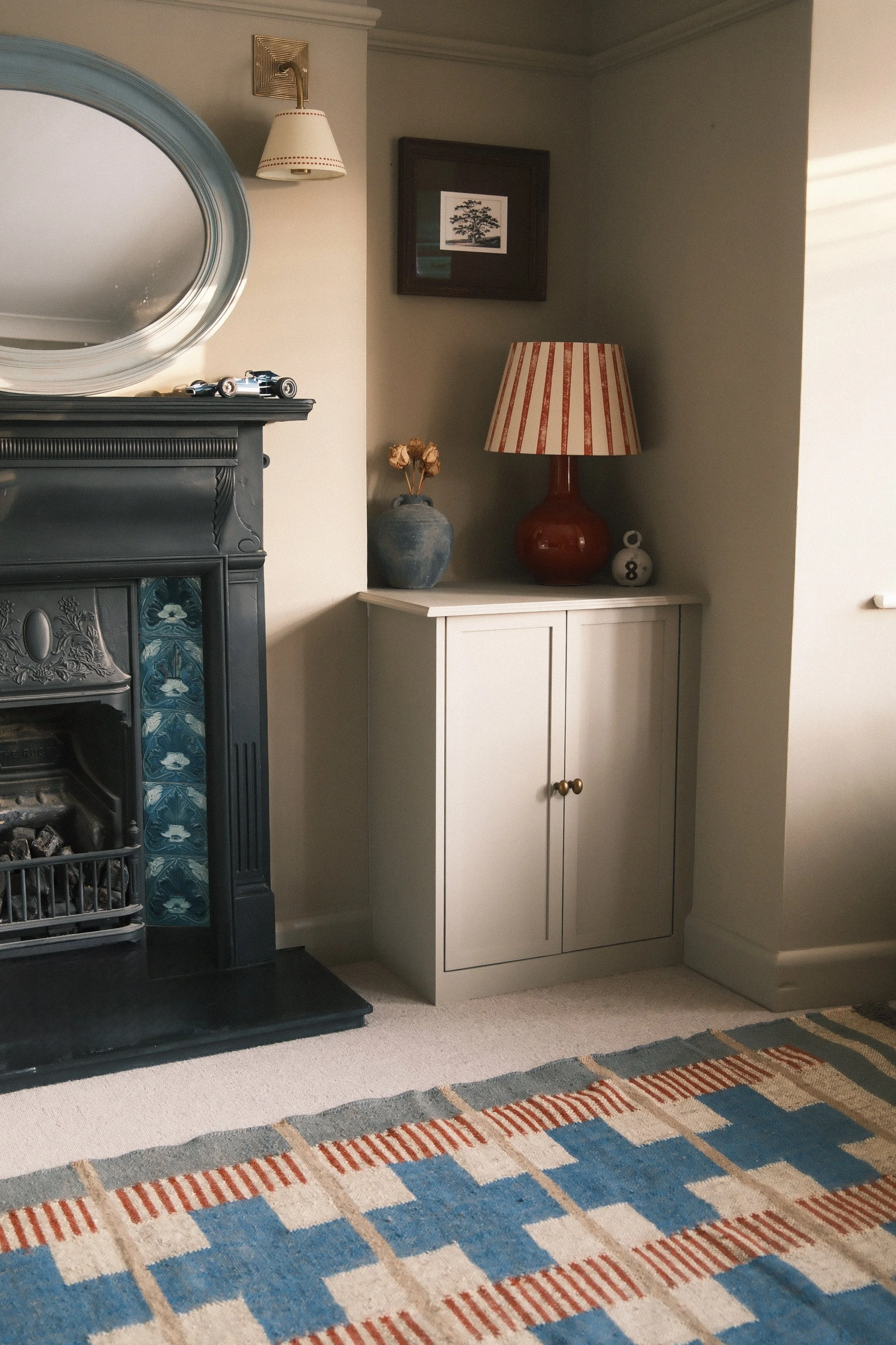 Interior of a living room with a fireplace, mirror, picture frame, table lamp, blue vase with dried flowers, and a patterned rug.