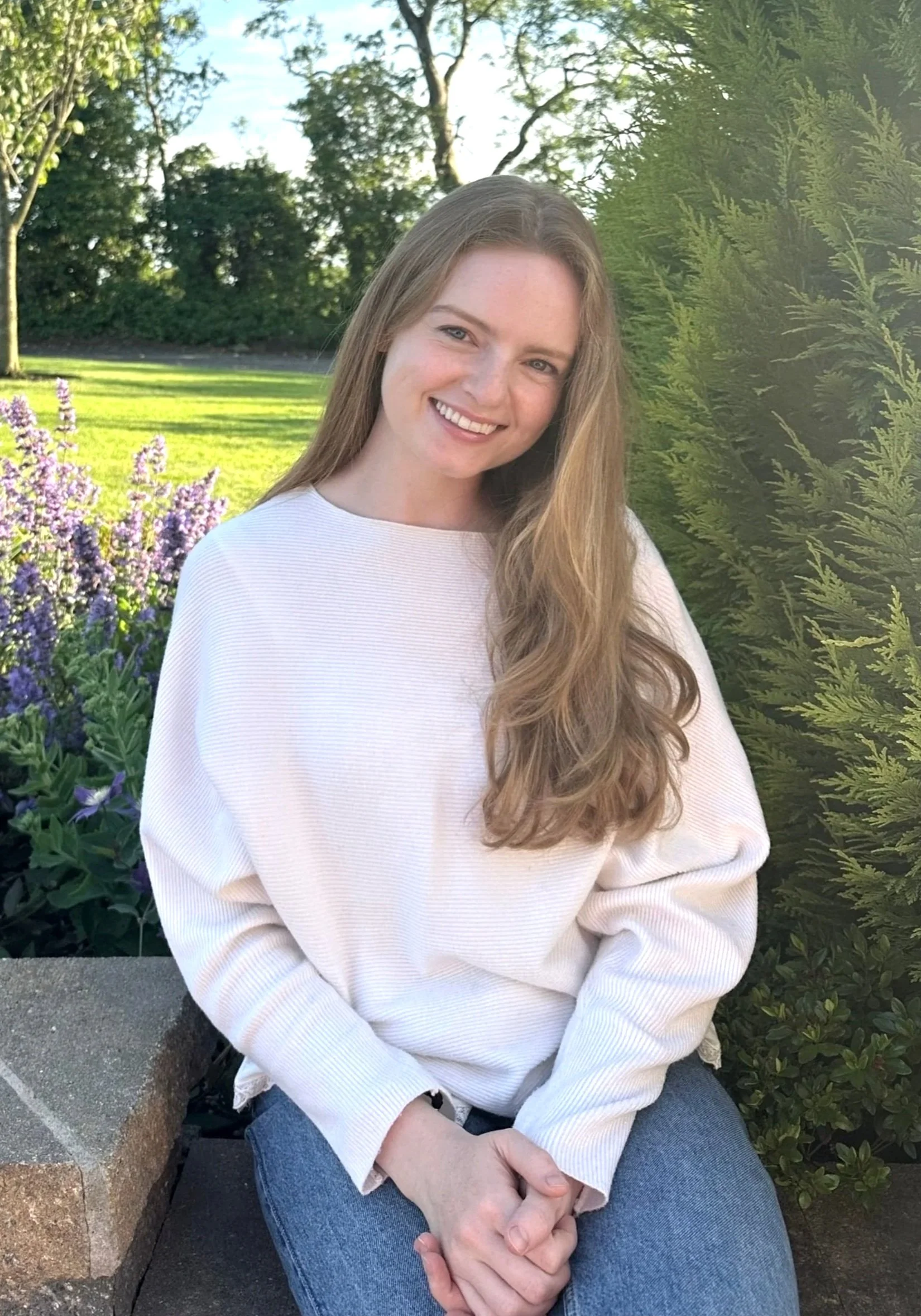 A young woman with long, wavy light brown hair smiling while sitting outdoors on a bench. She is wearing a white long-sleeve shirt and blue jeans. The background features green trees, a grassy area, and some purple flowers.