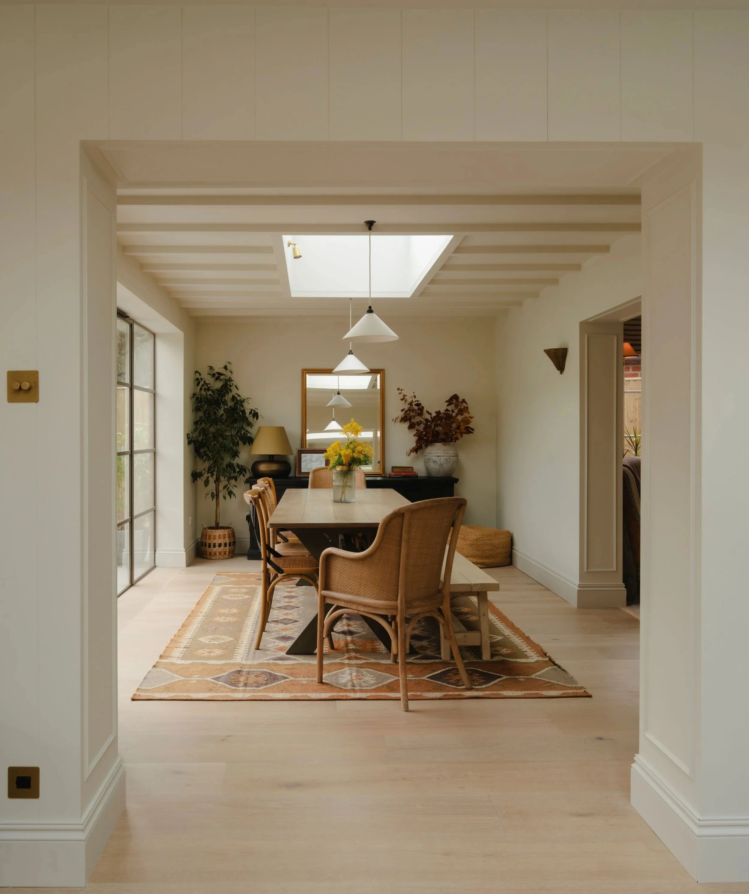 An open doorway leading to a dining room with a light wood table, surrounded by four wicker chairs. The room has a beige and tan geometric patterned rug, a window on the left side with natural light, and various plants and artwork, including a large mirror on the back wall.