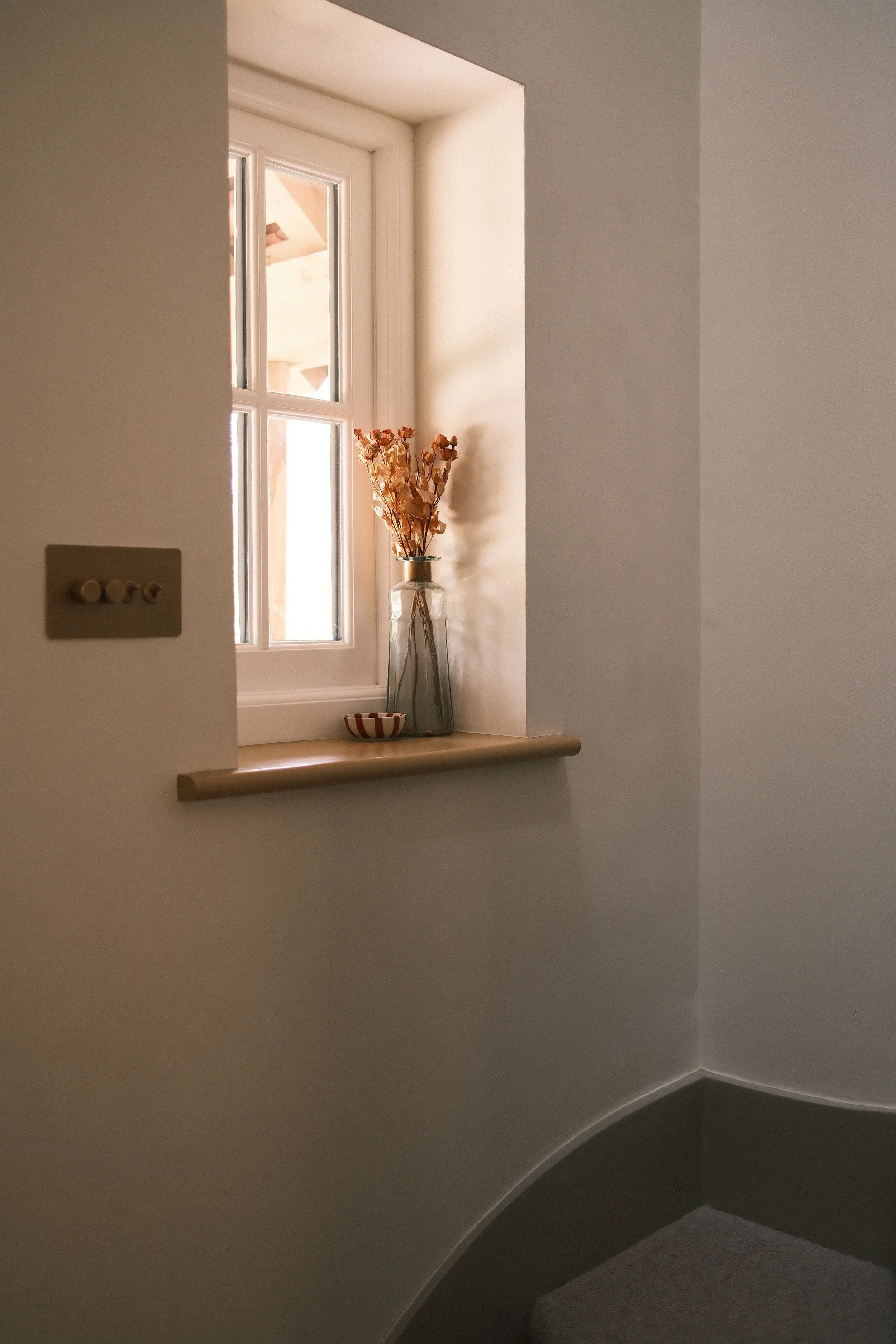 A windowsill with a glass vase holding dried flowers and a small striped bowl, with a window showing an outside view.