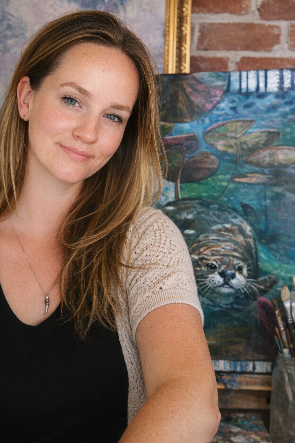A woman with long brown hair and blue eyes smiling, wearing a necklace and a beige knit sleeve, standing in front of a colorful painting of lily pads and a cat peeking out from water.