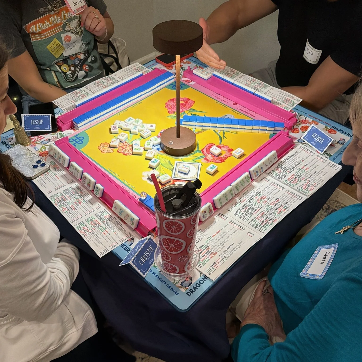 A group of people playing a board game called Mahjong around a table, with a pink and yellow game set, tiles, score sheets, and drinks on the table.