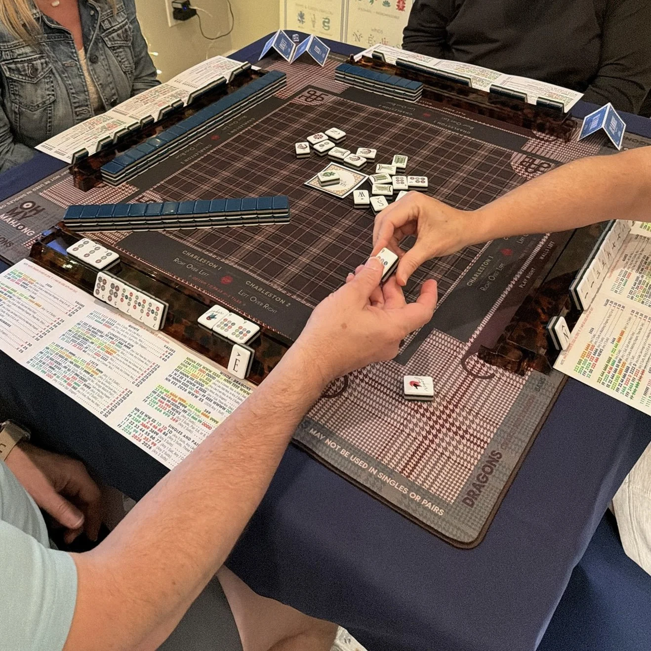 A group of people playing dominoes on a table with betting sheets and scorecards around the game's edges.