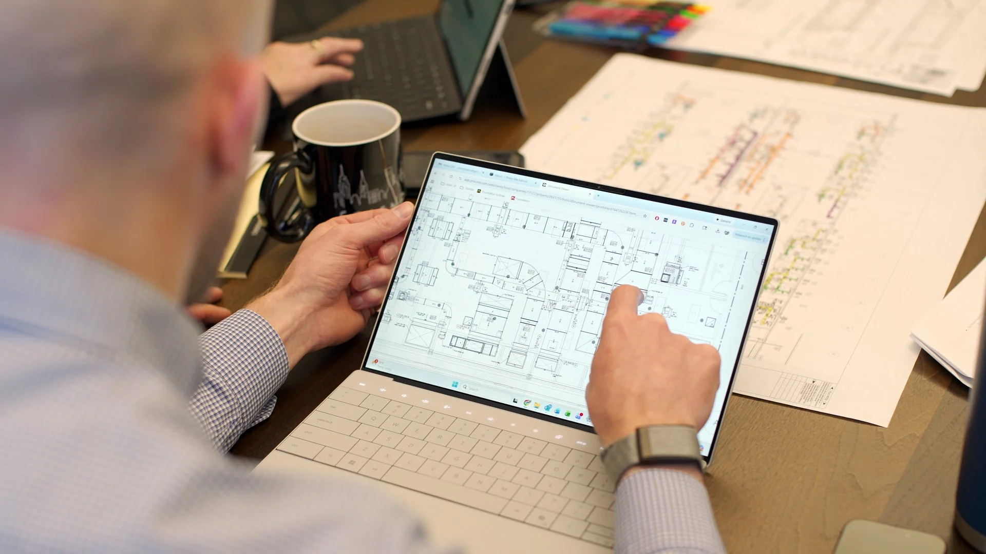 Person holding a tablet displaying an architectural floor plan, with architectural drawings and colored markers on the table.
