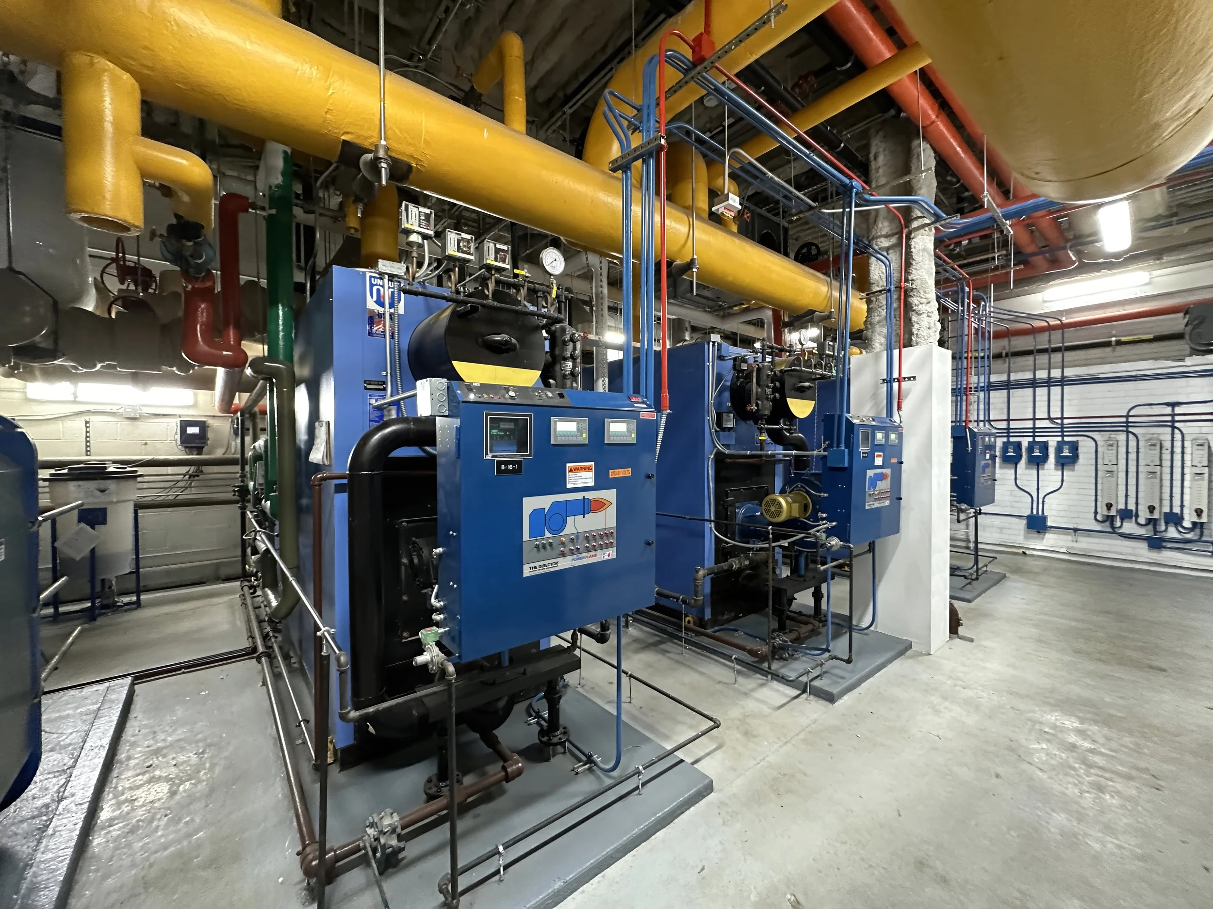 Image of industrial equipment, specifically a boiler system with blue machinery, pipes, and gauges in a mechanical room.