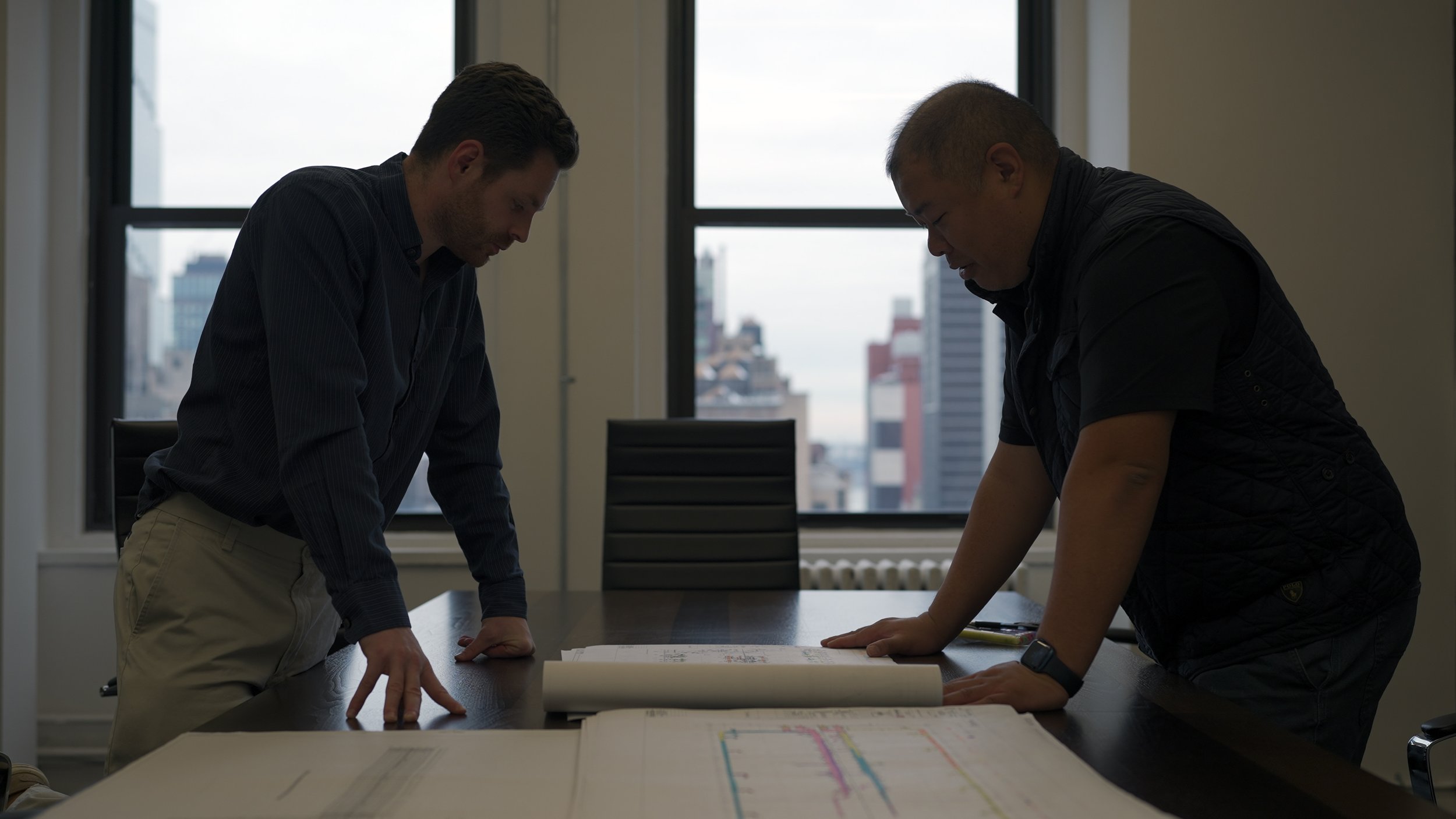 Two men looking down at papers and blueprints on a conference table in a bright office with large windows showing a city skyline.