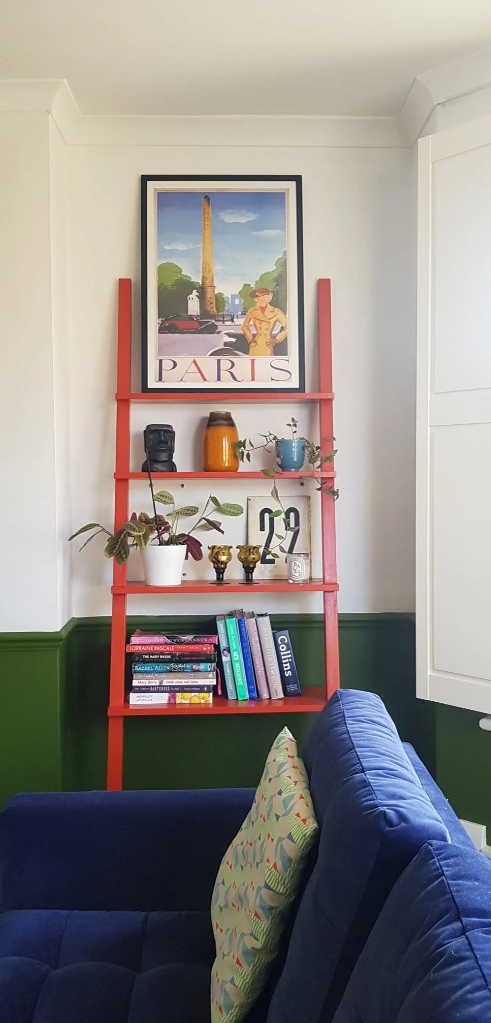 A red ladder-style bookshelf with a framed Paris poster on top, various decorative items, a plant, books, and two small gold-colored goblets. Part of a blue sofa with a patterned pillow is in the foreground.
