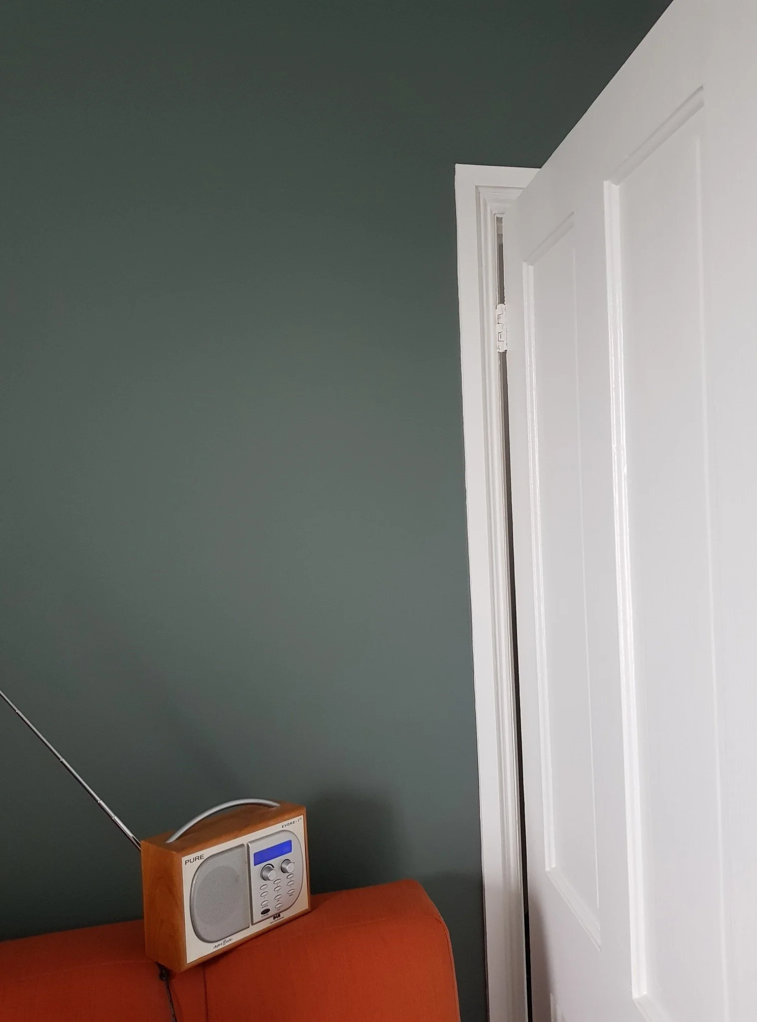 A room corner with gray wall, white door ajar, orange couch, and a vintage wooden radio with a blue digital display and antenna.