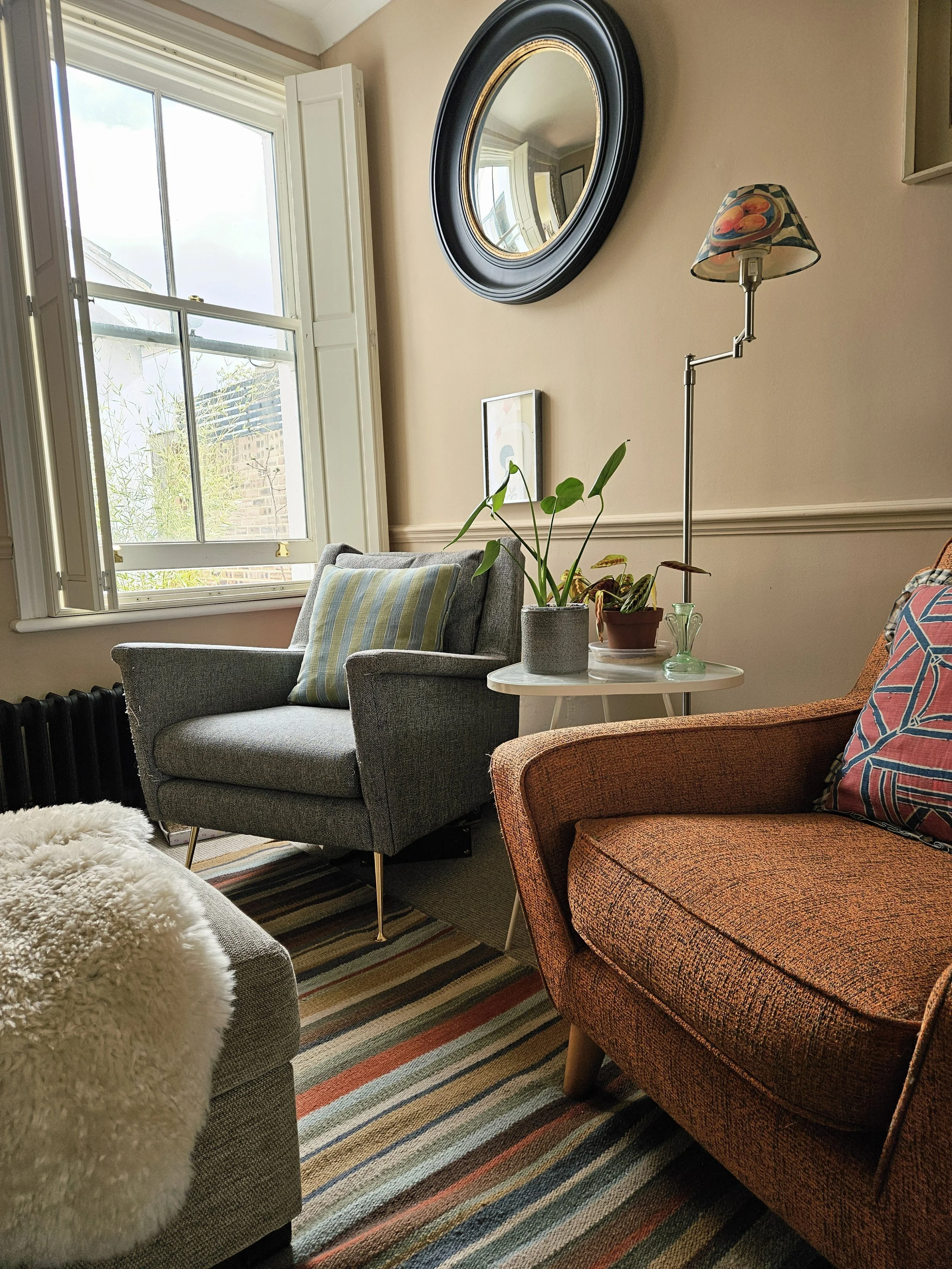 A cozy living room corner with a window, a gray armchair with a striped pillow, a small white side table with potted plants and a small vase, an orange armchair with a patterned pillow, a black radiator, a rainbow-striped rug, a wall mirror, and a floor lamp with a colorful lampshade.