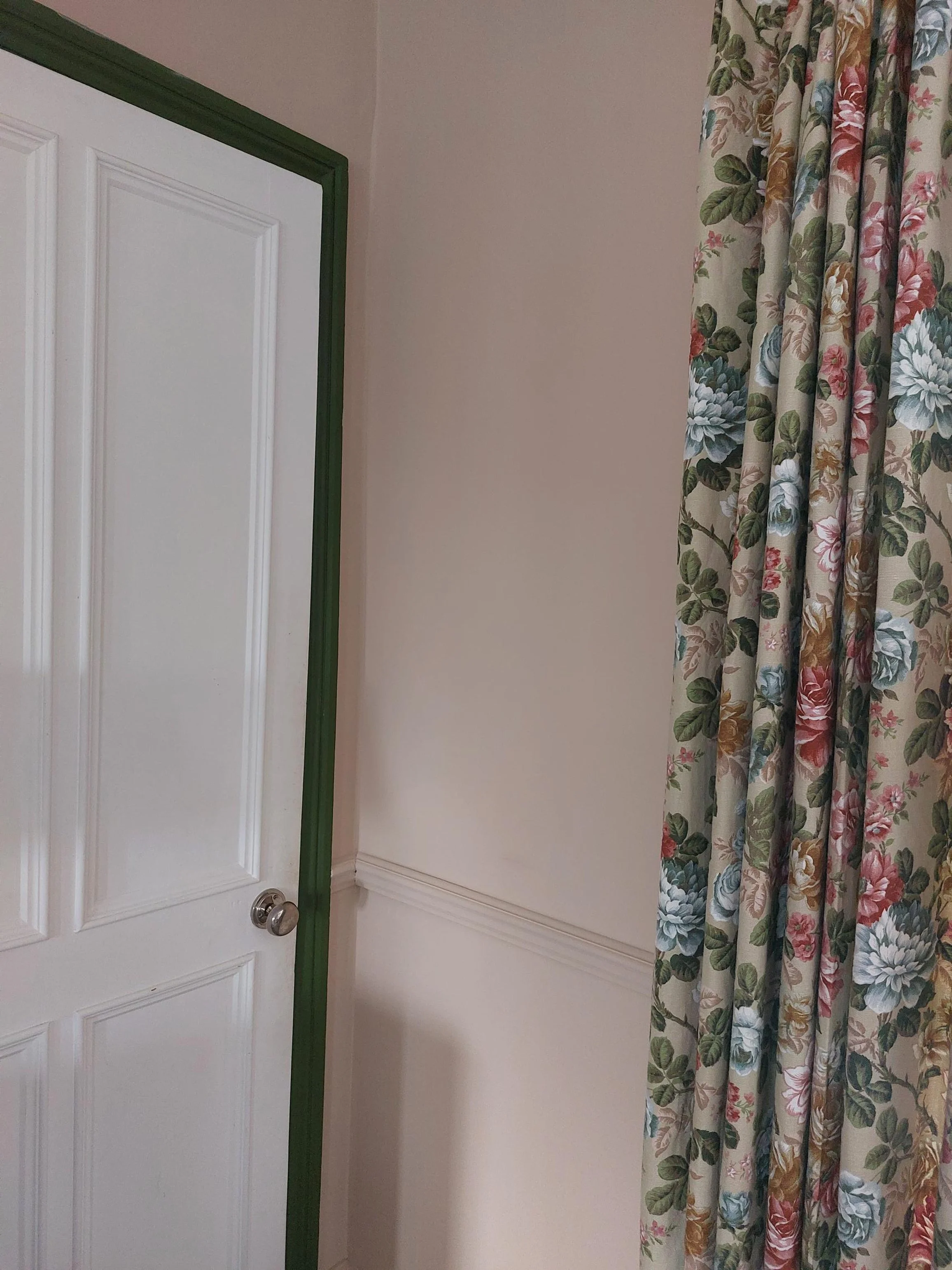 Interior corner of a room featuring a white door with green trim and a floral curtain with green, pink, blue, and cream-colored flowers and leaves.