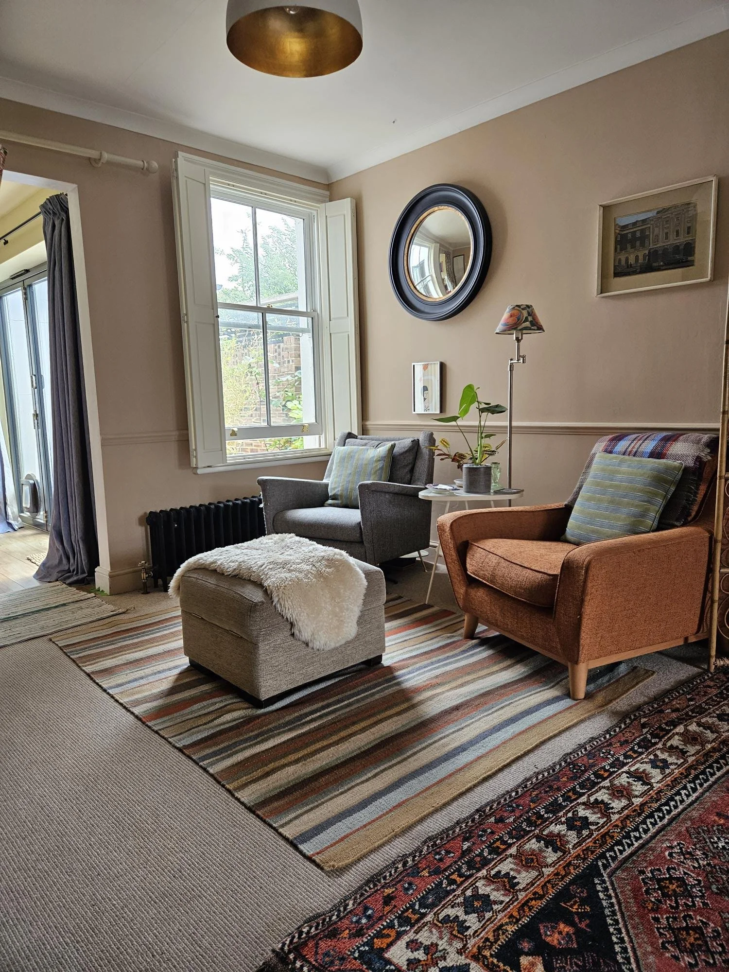 Cozy living room with beige walls, a window with white shutters, a gray armchair with striped pillows, a small ottoman with a fluffy cover, a colorful rug, a standing lamp with a decorative lampshade, and framed artwork on the wall.