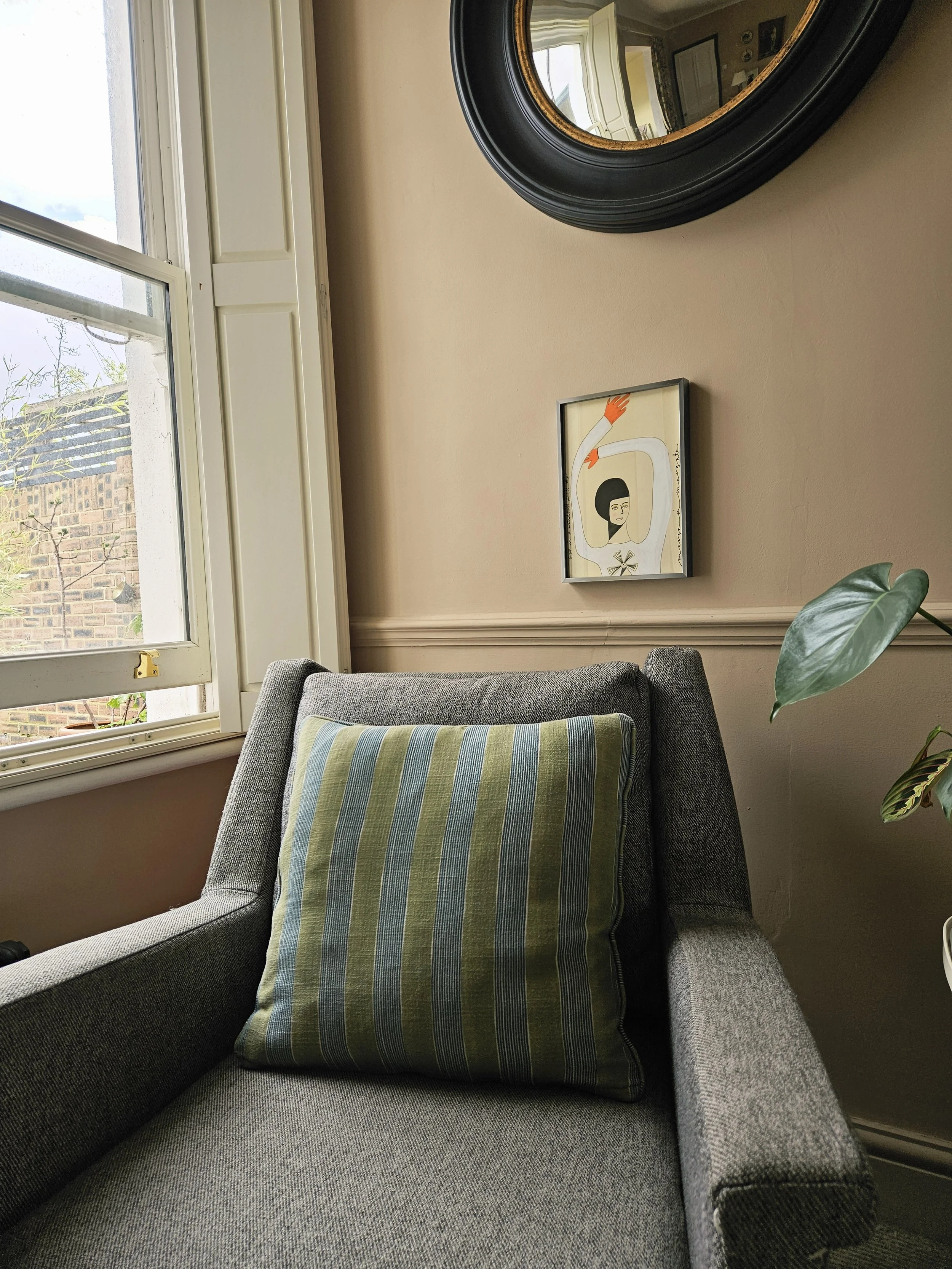 A cozy corner with a gray armchair, a striped green and blue pillow, a round black mirror on the wall, a framed abstract artwork, a large window with white trim, and a potted plant with large green leaves.