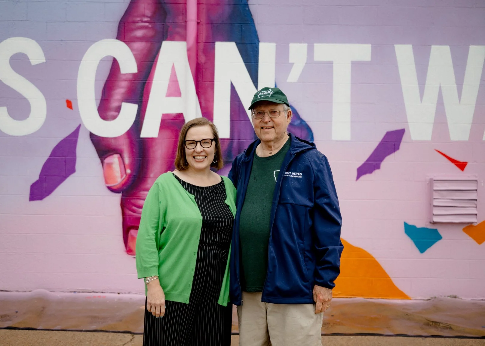 Two people smiling together in front of the mural.