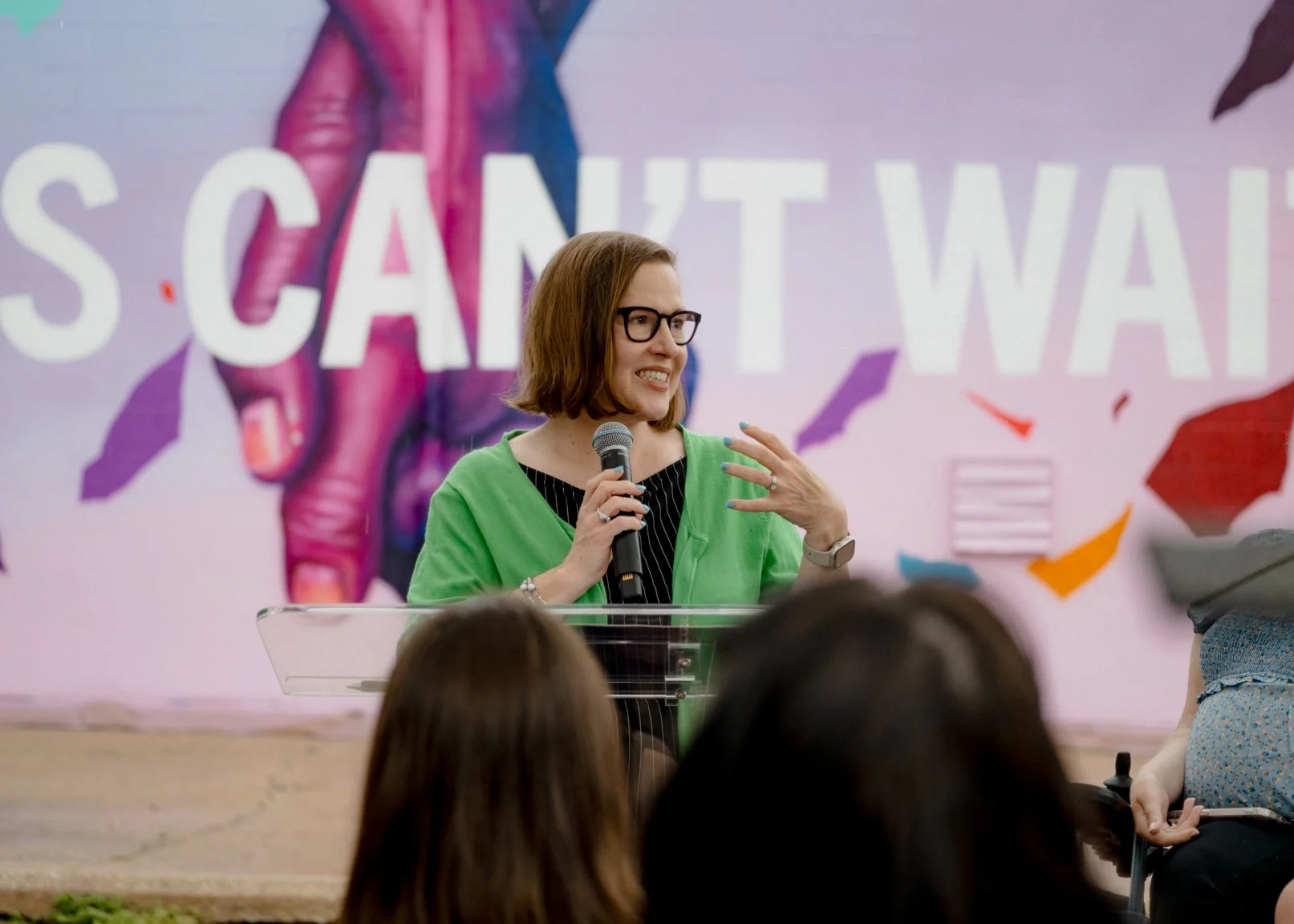 A woman in green speaking into a microphone in front of the mural.