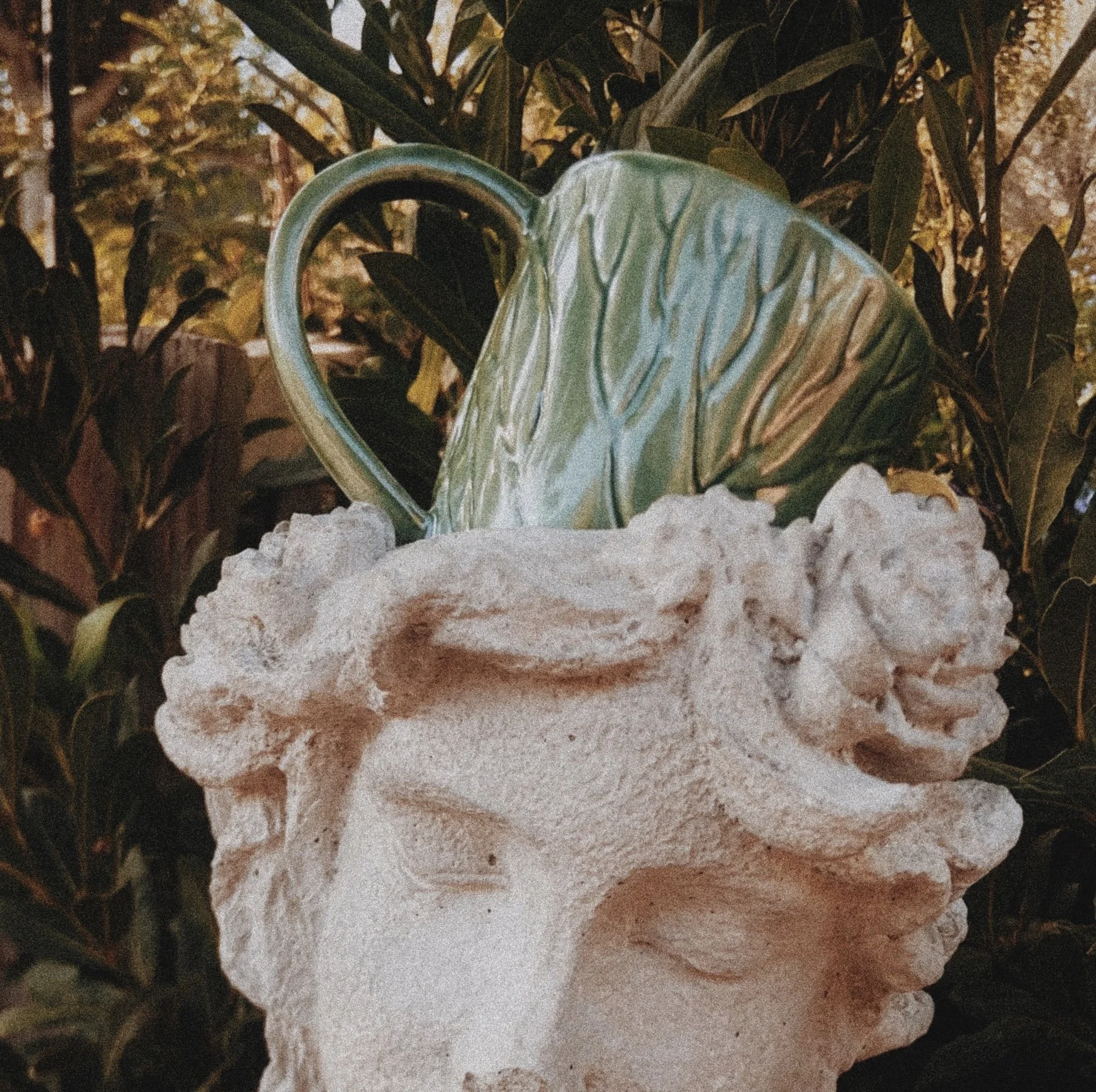 A decorative ceramic mug with green leaf patterns atop a stone sculpture of a lion's head, surrounded by green foliage.