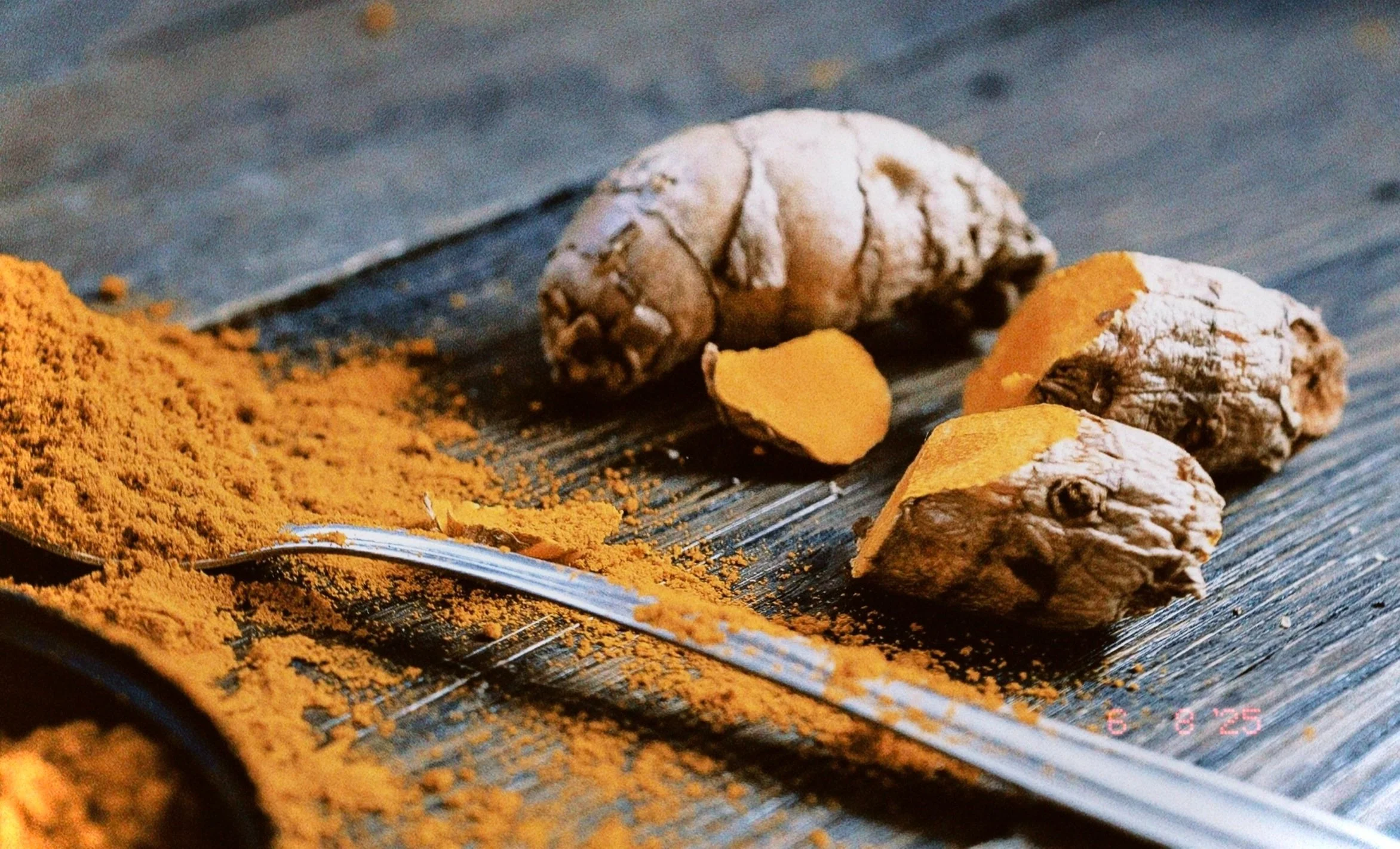 Three pieces of fresh turmeric root, some of which are sliced, on a dark wooden surface next to a pile of ground turmeric spice and a spoon.