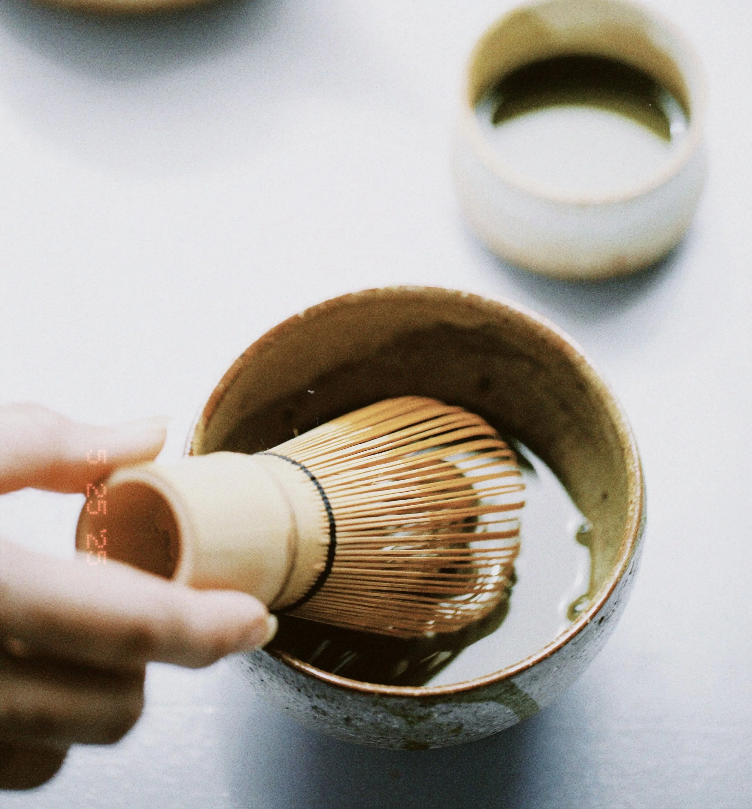 Person using a bamboo whisk to prepare matcha tea in a ceramic bowl on a white surface, with a cup of green tea in the background.