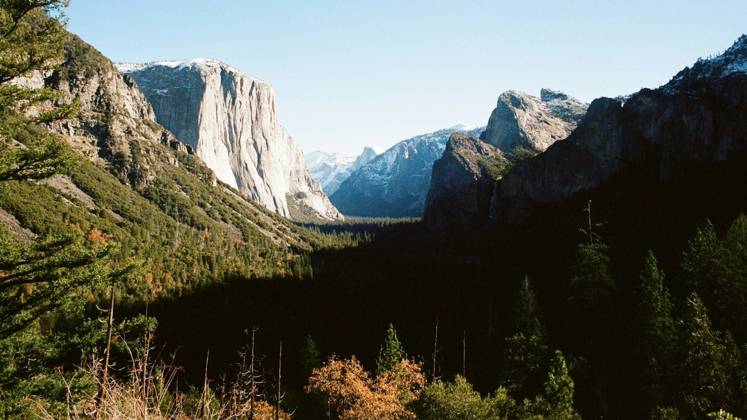 A scenic view of a mountainous landscape with tall granite cliffs and snow-capped peaks in the background, surrounded by dense green pine forests.