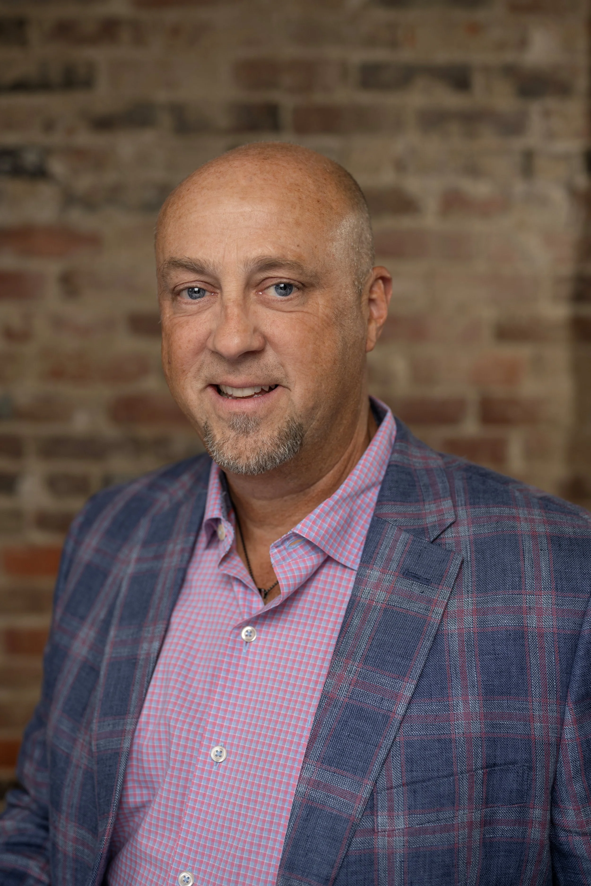 A man with a bald head, beard, and blue eyes wearing a checkered shirt and blazer with a brick wall background.