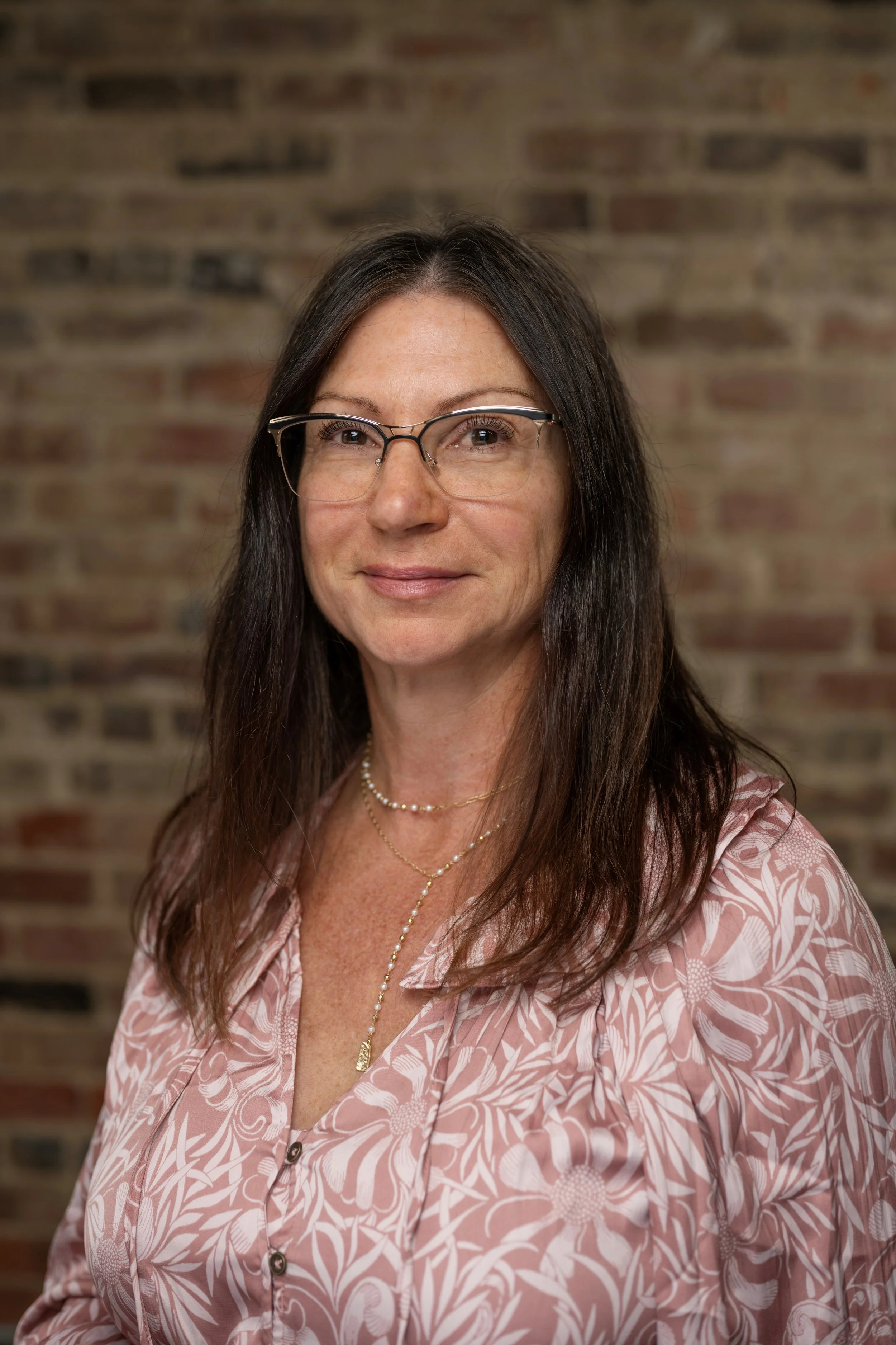 Portrait of a woman with long dark hair, glasses, a pink floral blouse, and layered necklaces, standing in front of a brick wall.