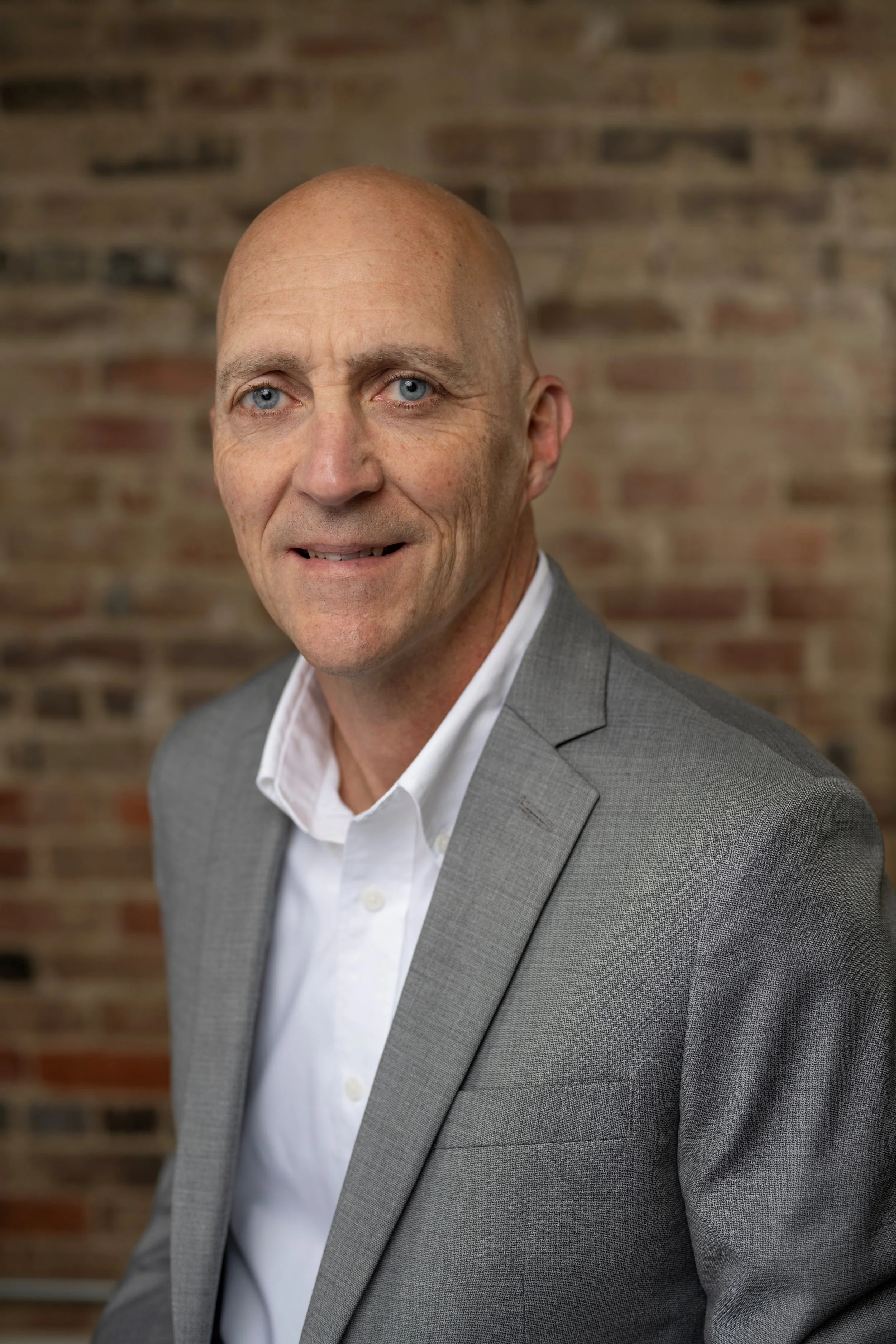 A middle-aged man with a bald head and blue eyes wearing a light gray suit jacket and white shirt, standing against a brick wall.