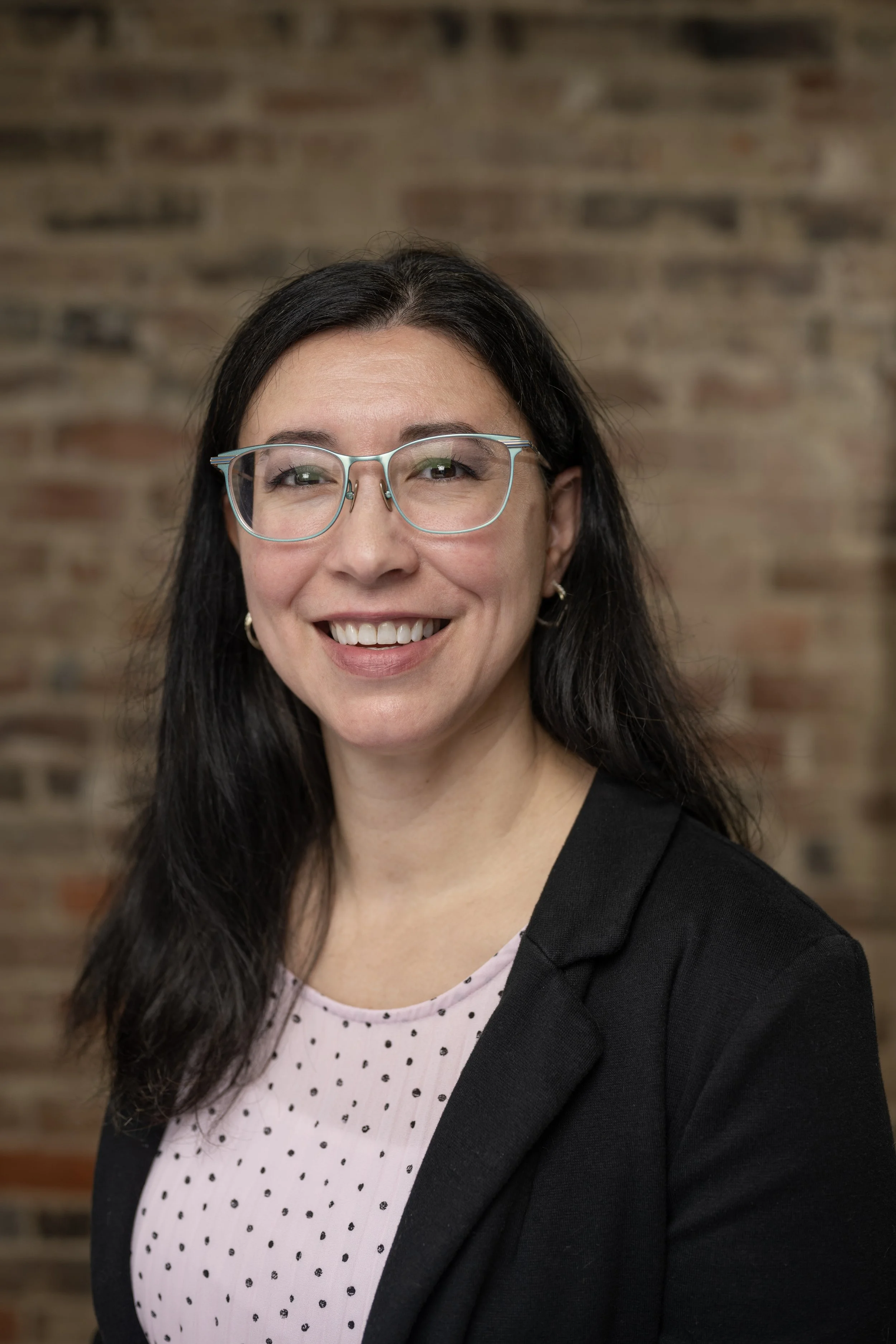 A woman with long dark hair and glasses smiling, wearing a black blazer and pink polka-dot top, standing against a brick wall.