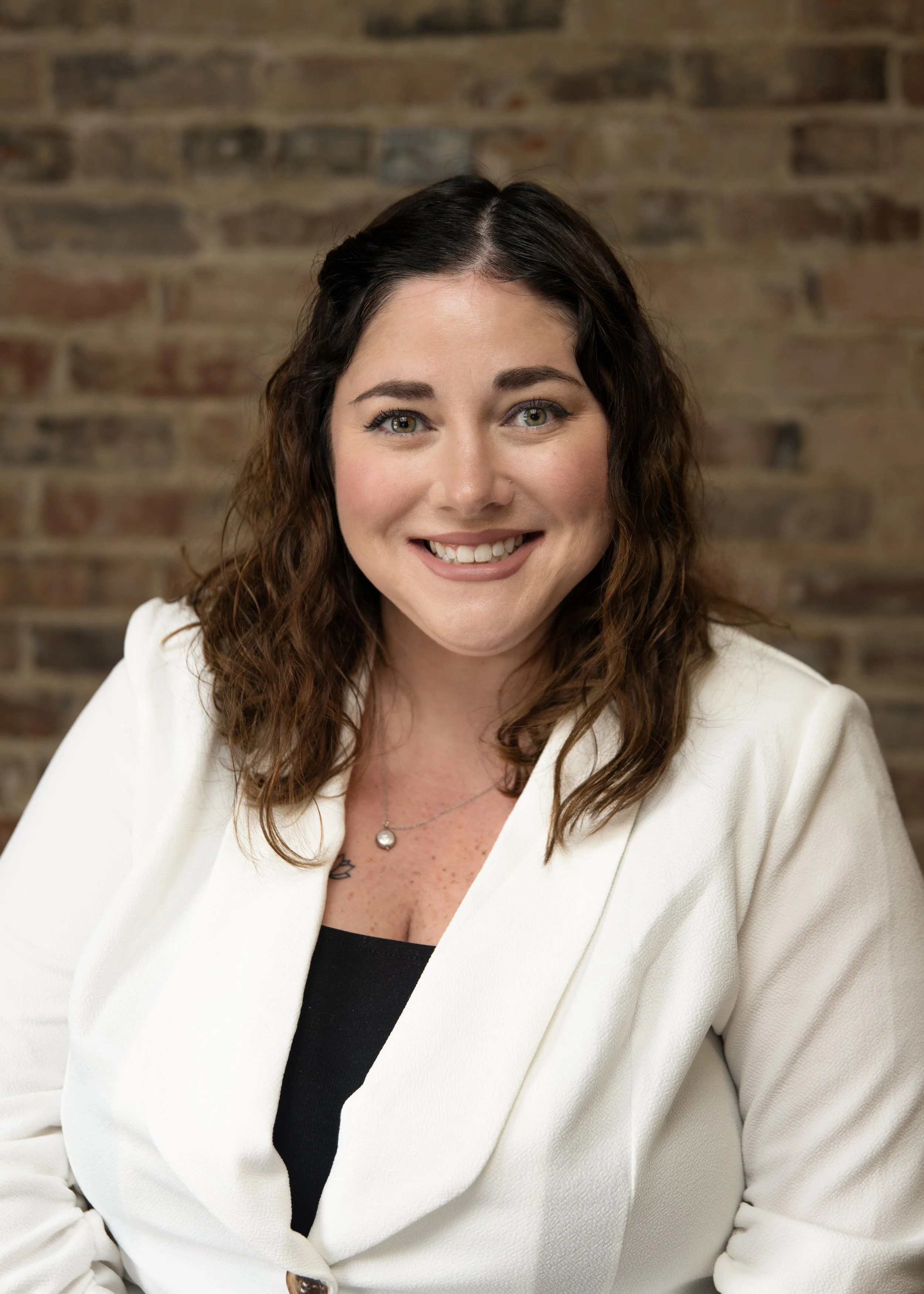 A smiling woman with curly brown hair, wearing a white blazer and black top, posing against a brick wall background.