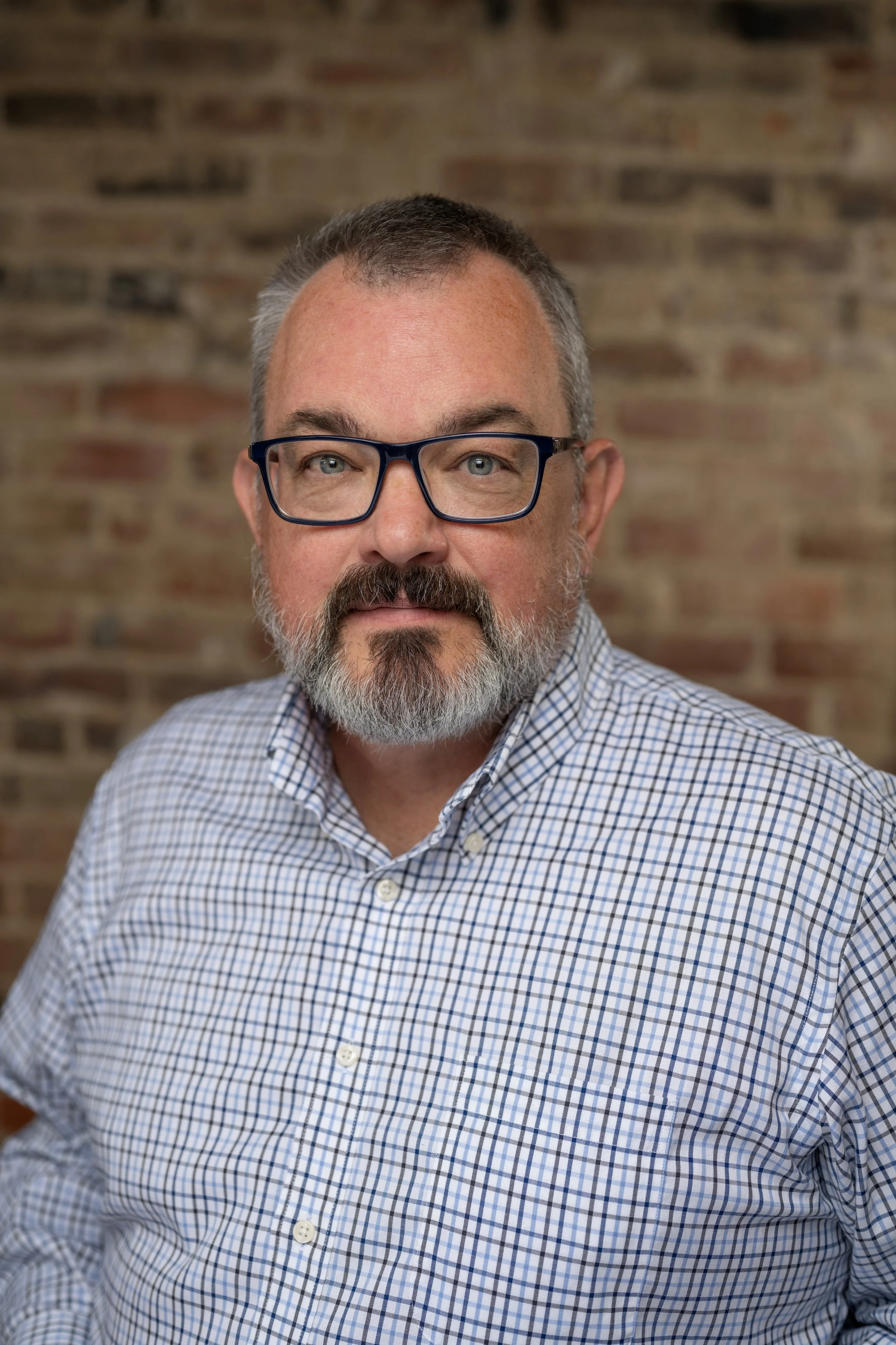 A middle-aged man with short gray hair, a beard, and glasses, wearing a checkered shirt, standing in front of a brick wall.