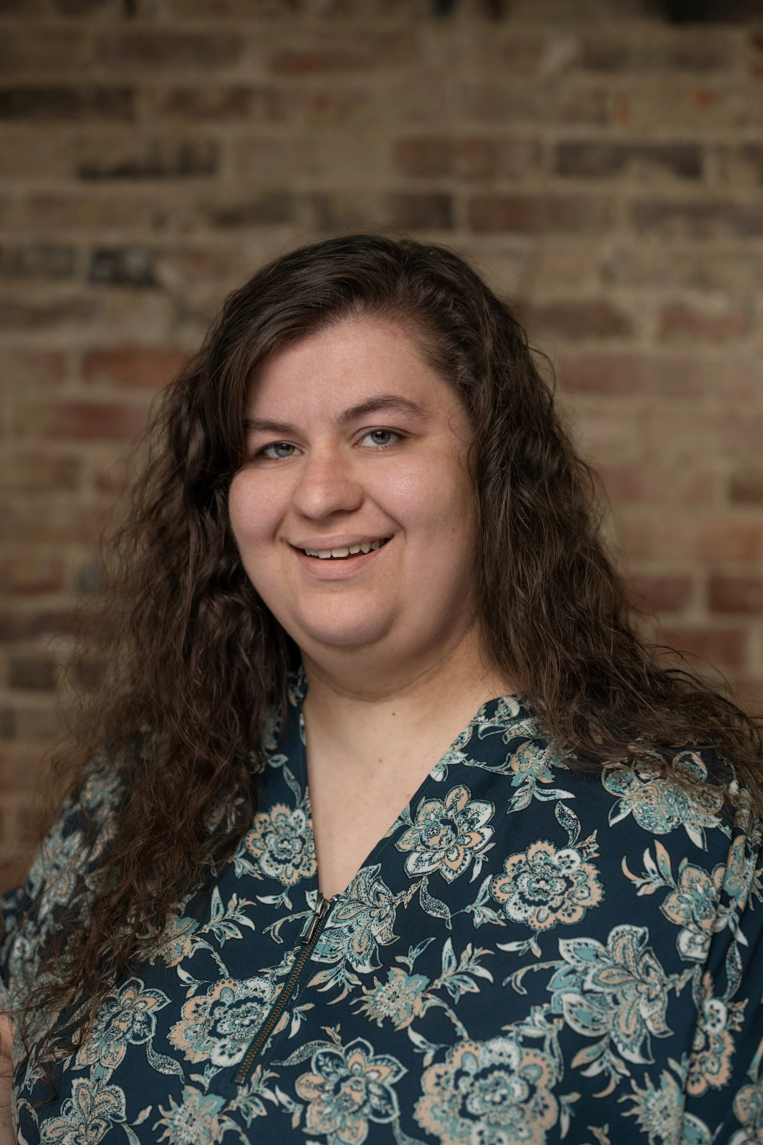 A woman with long, curly brown hair, blue eyes, and a fair complexion, smiling at the camera, wearing a navy blue floral blouse with a zipper, standing in front of a brick wall.