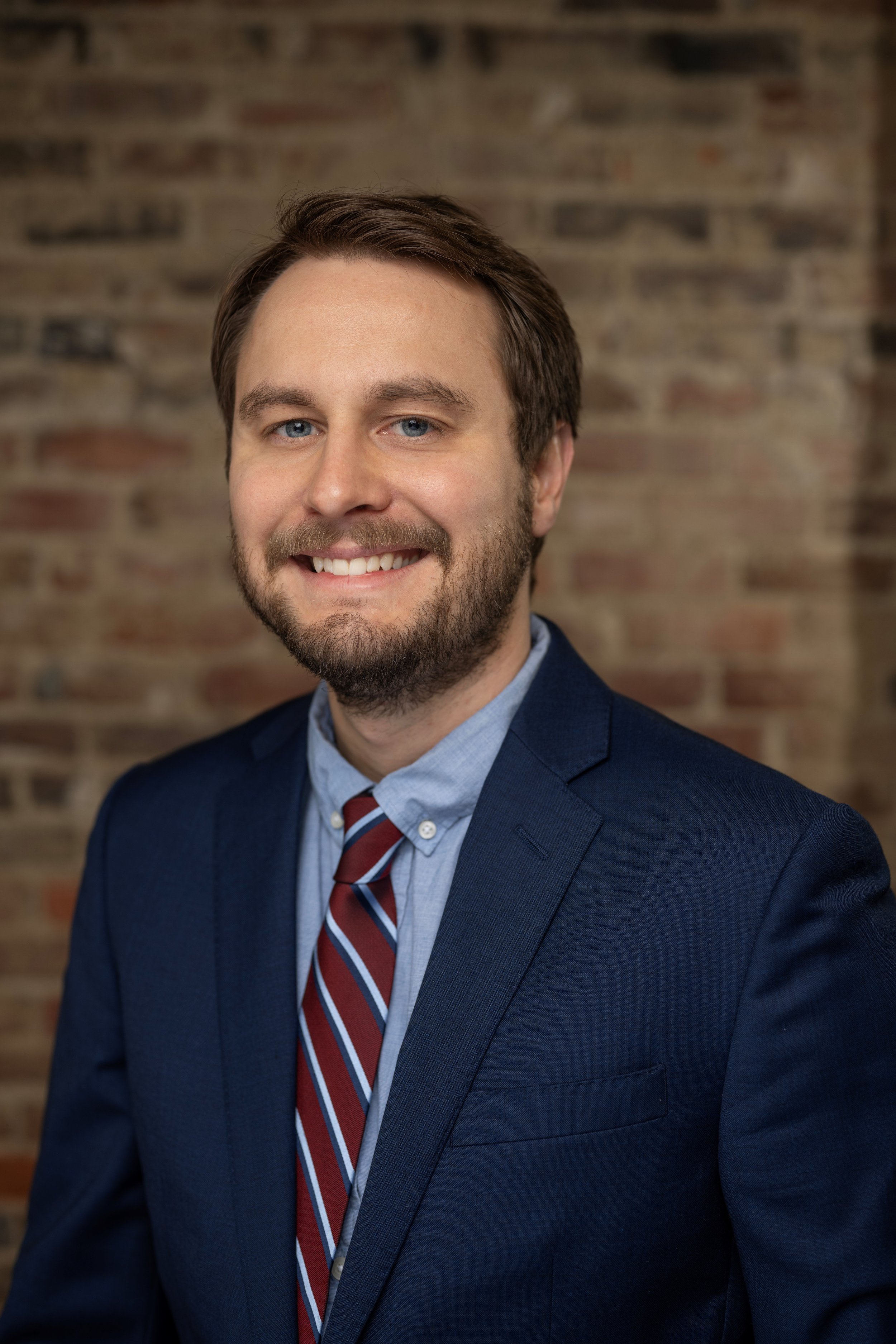 A man with short brown hair, blue eyes, and a beard, wearing a navy suit, light blue shirt, and red striped tie, smiling in front of a brick wall.