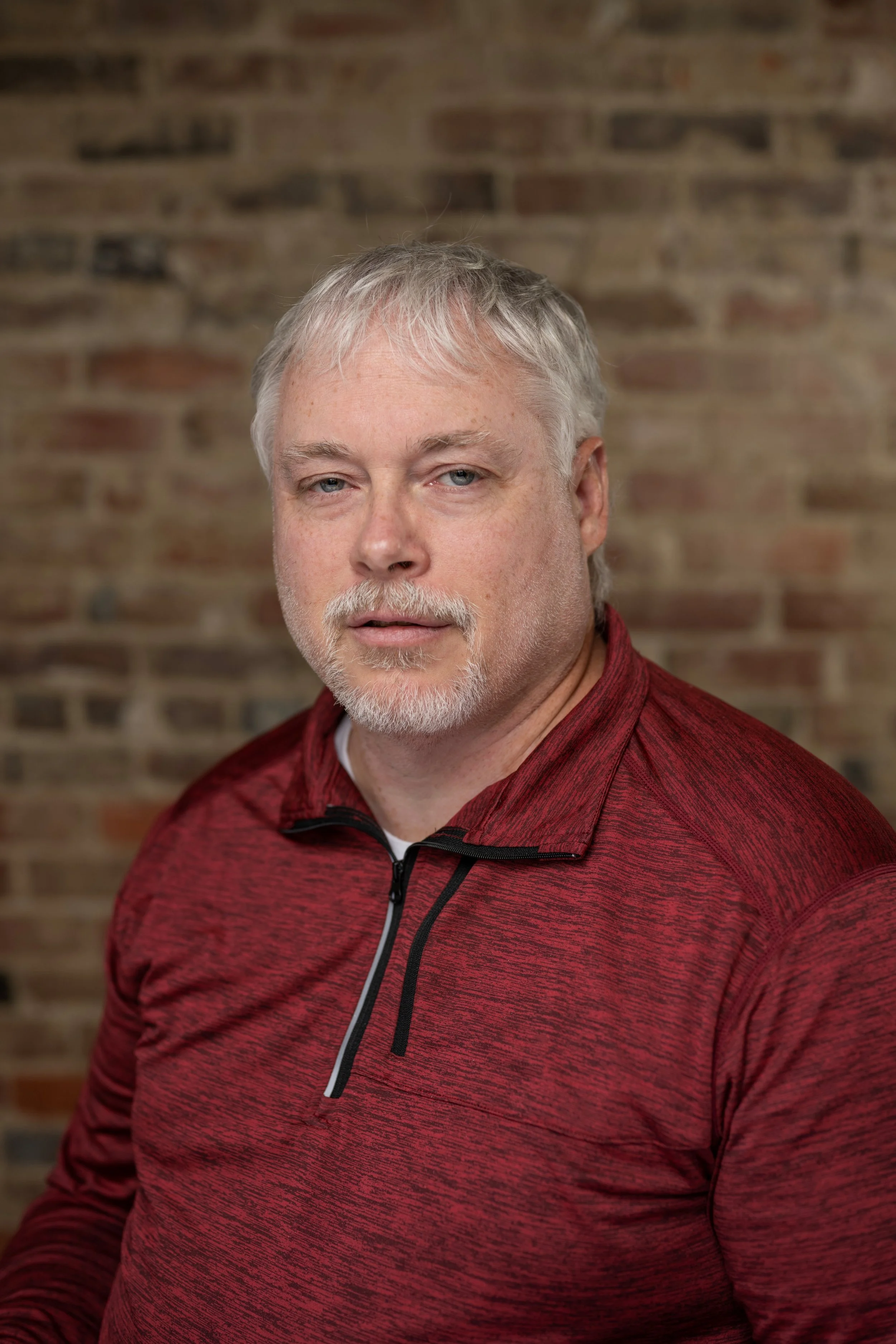 A middle-aged man with gray hair and beard, wearing a red athletic jacket, standing against a brick wall background.