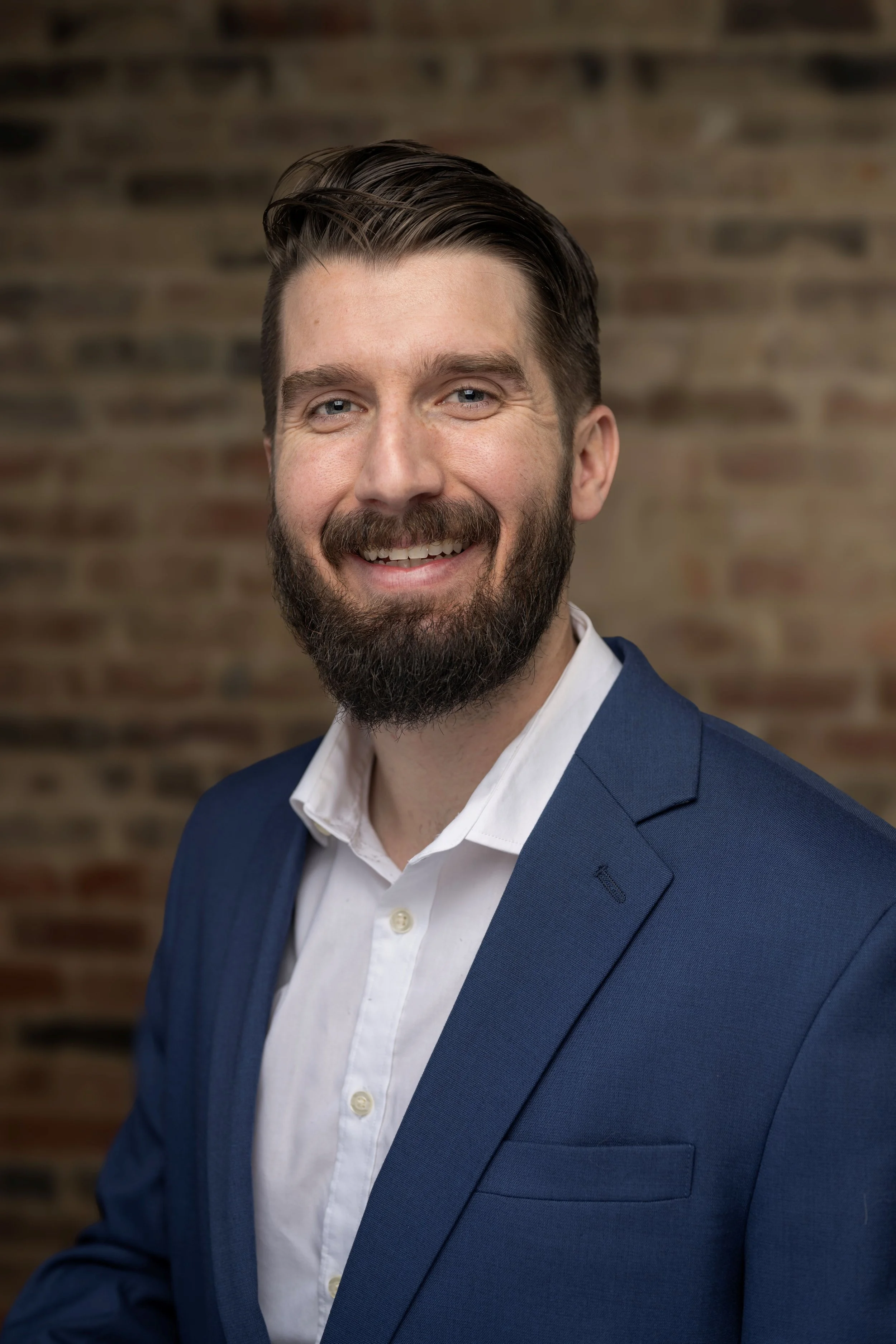 A smiling man with a beard wearing a navy blue suit jacket and white shirt against a brick wall background.
