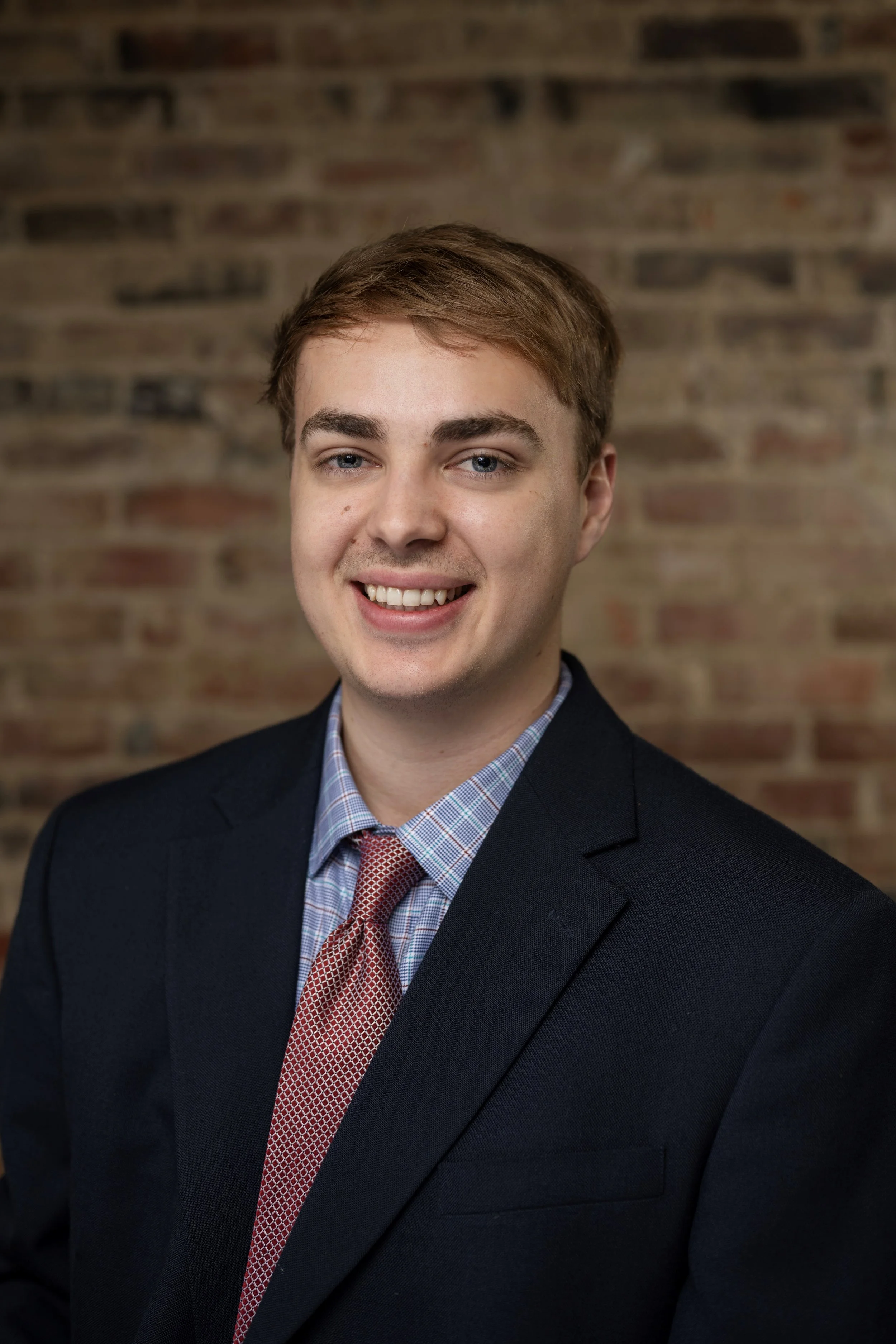 A young man with light brown hair, blue eyes, and a fair complexion, smiling, wearing a dark suit, checkered shirt, and a red tie, standing against a blurred brick wall background.