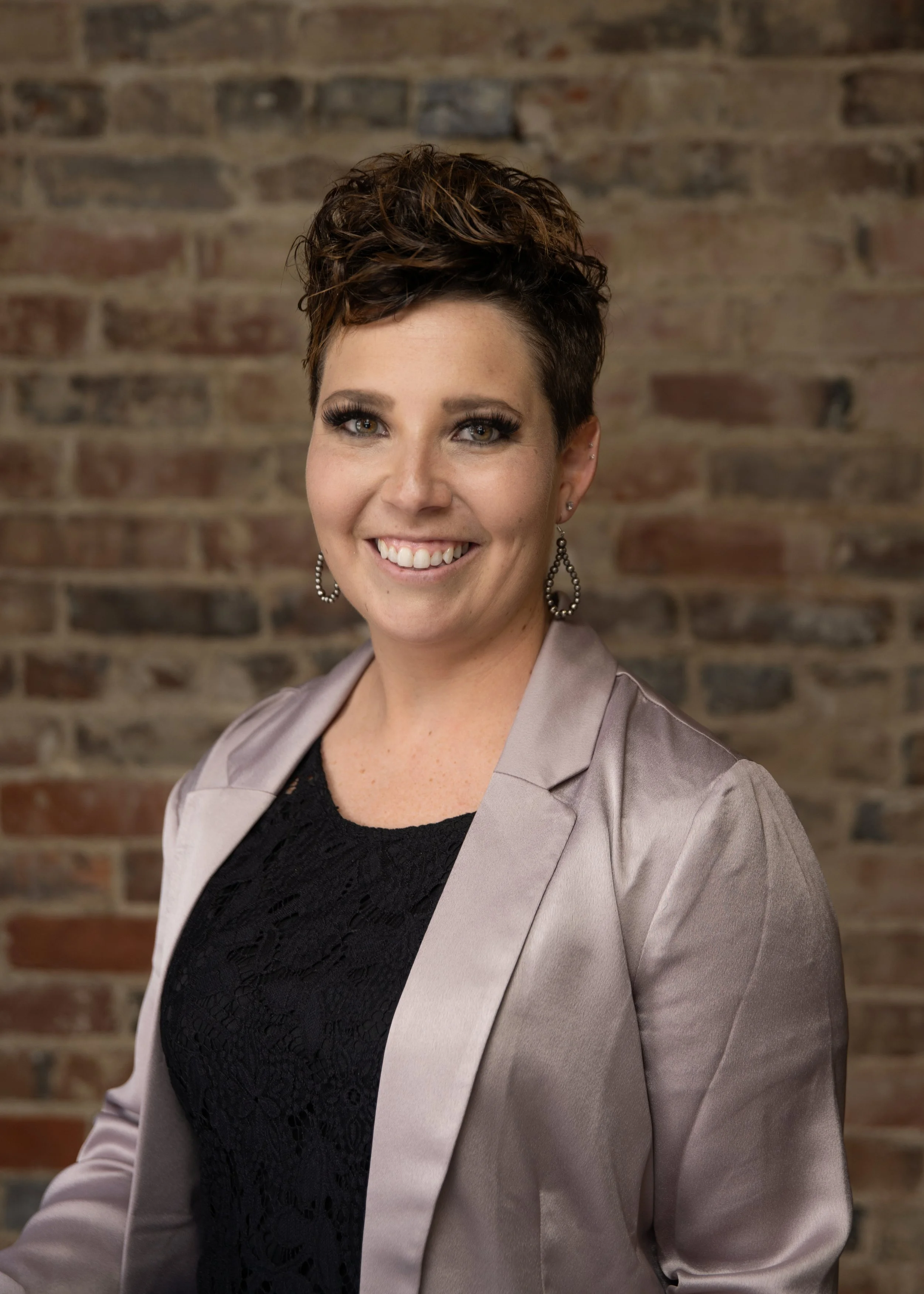 A woman smiling at the camera with short, styled hair, wearing a black lace top, a beige satin blazer, and silver earrings against a brick wall background.